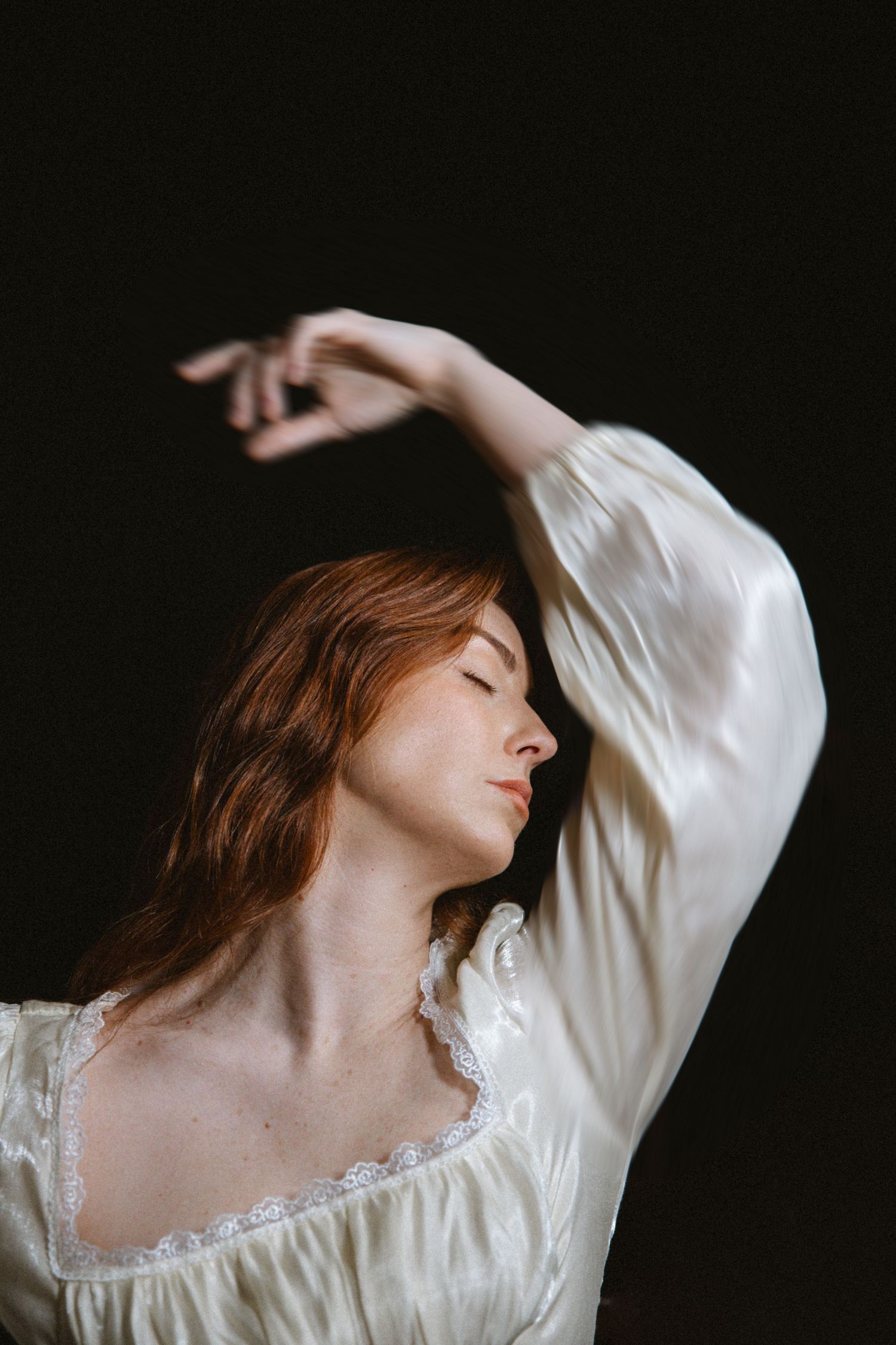 A woman in a vintage white dress with lace trim poses gracefully against a dark background in an artistic portrait series.