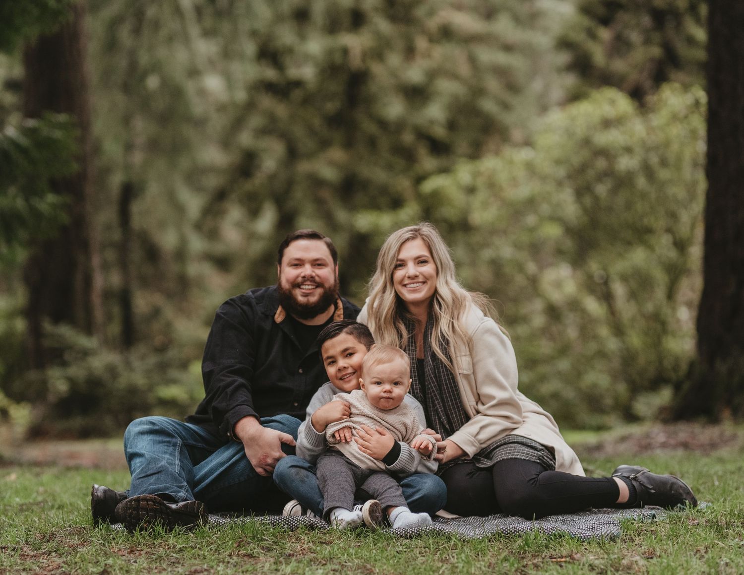 Family sitting together outdoors on grass with evergreen forest background.