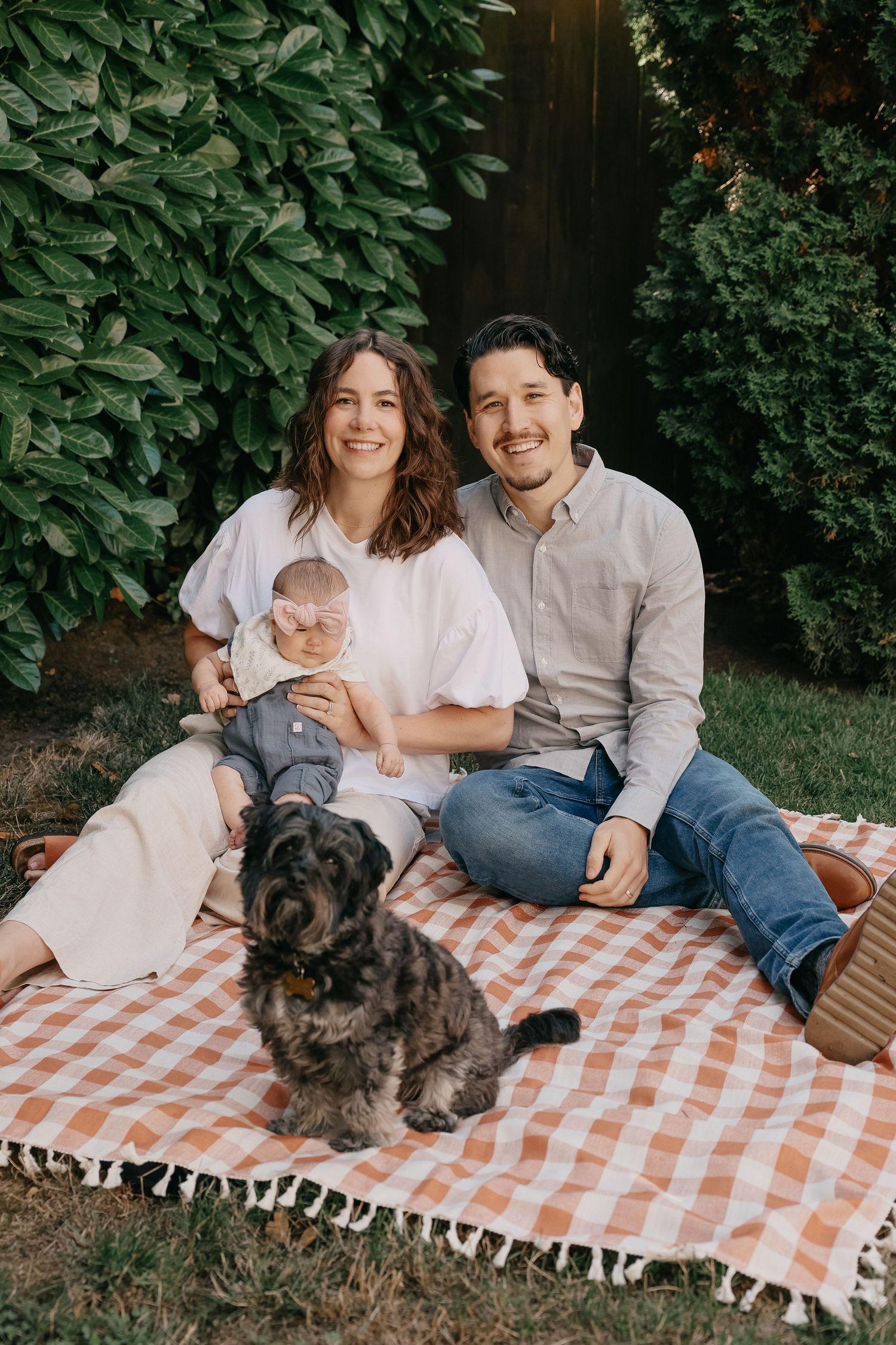 Family picnic scene with dog on checkered blanket in leafy backyard setting.