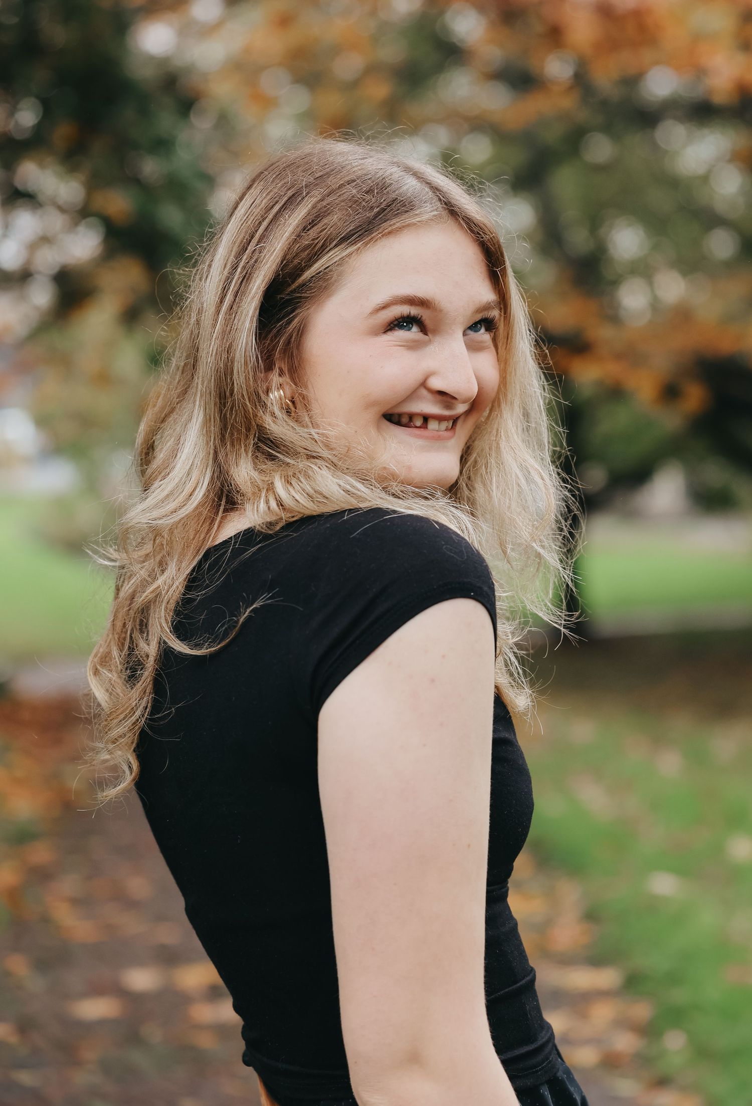 Person in black top smiling while looking over shoulder in autumn park setting.