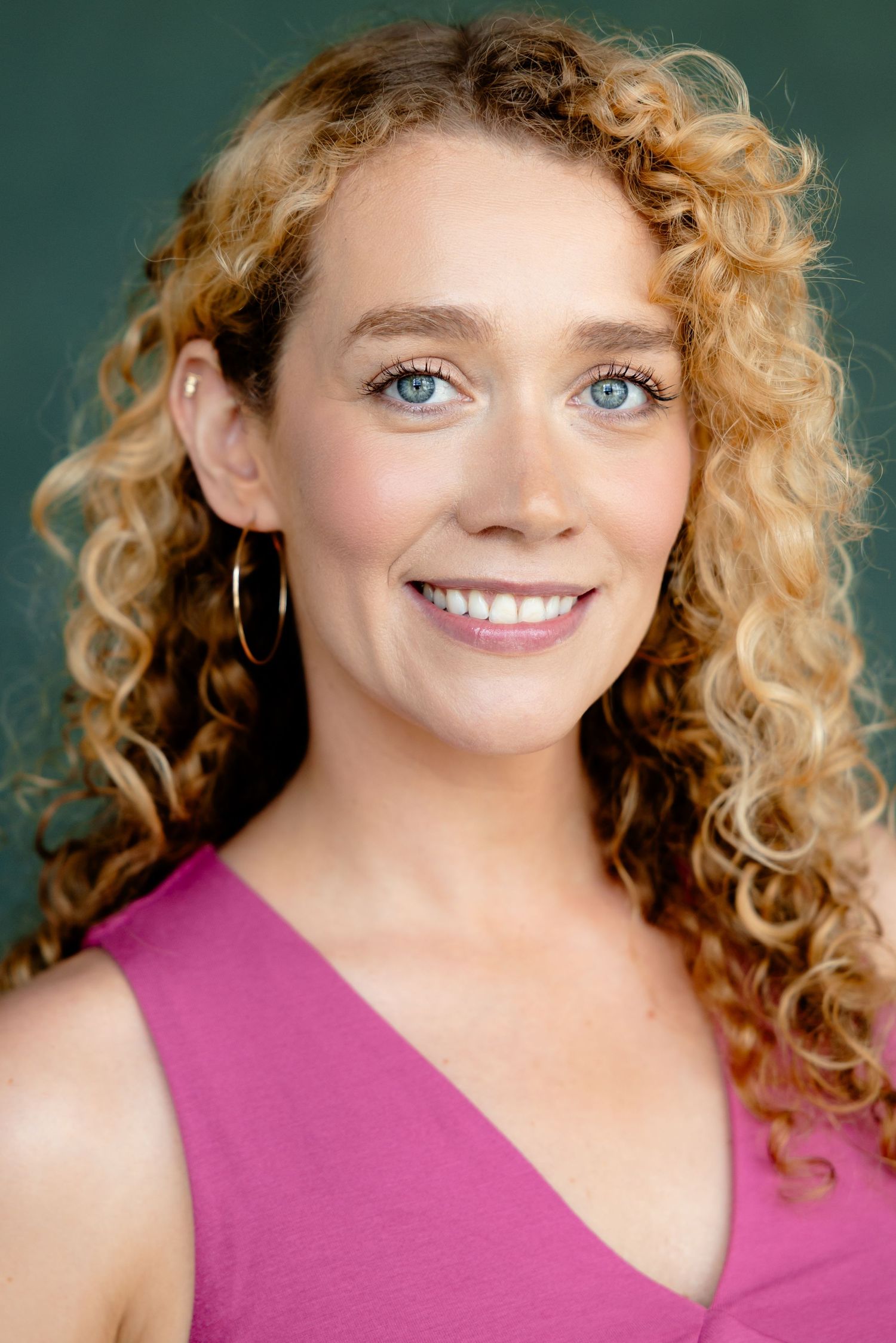 A smiling woman with curly blonde hair and blue eyes wearing a pink top and gold hoop earrings against a green background.