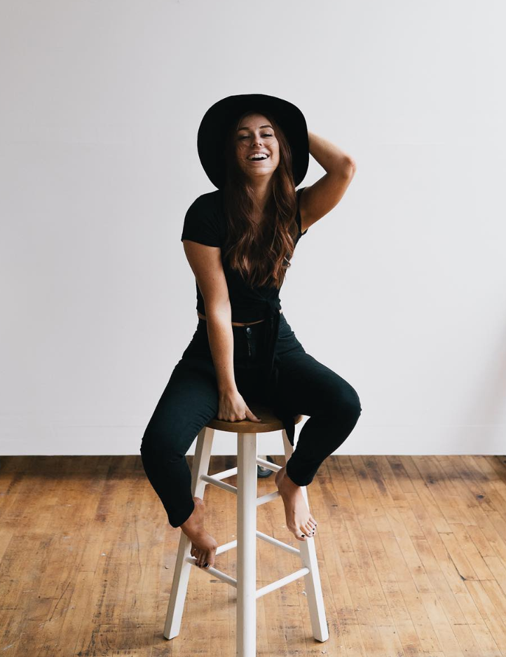 Joyful portrait of person in black outfit and hat sitting on wooden stool against white background.