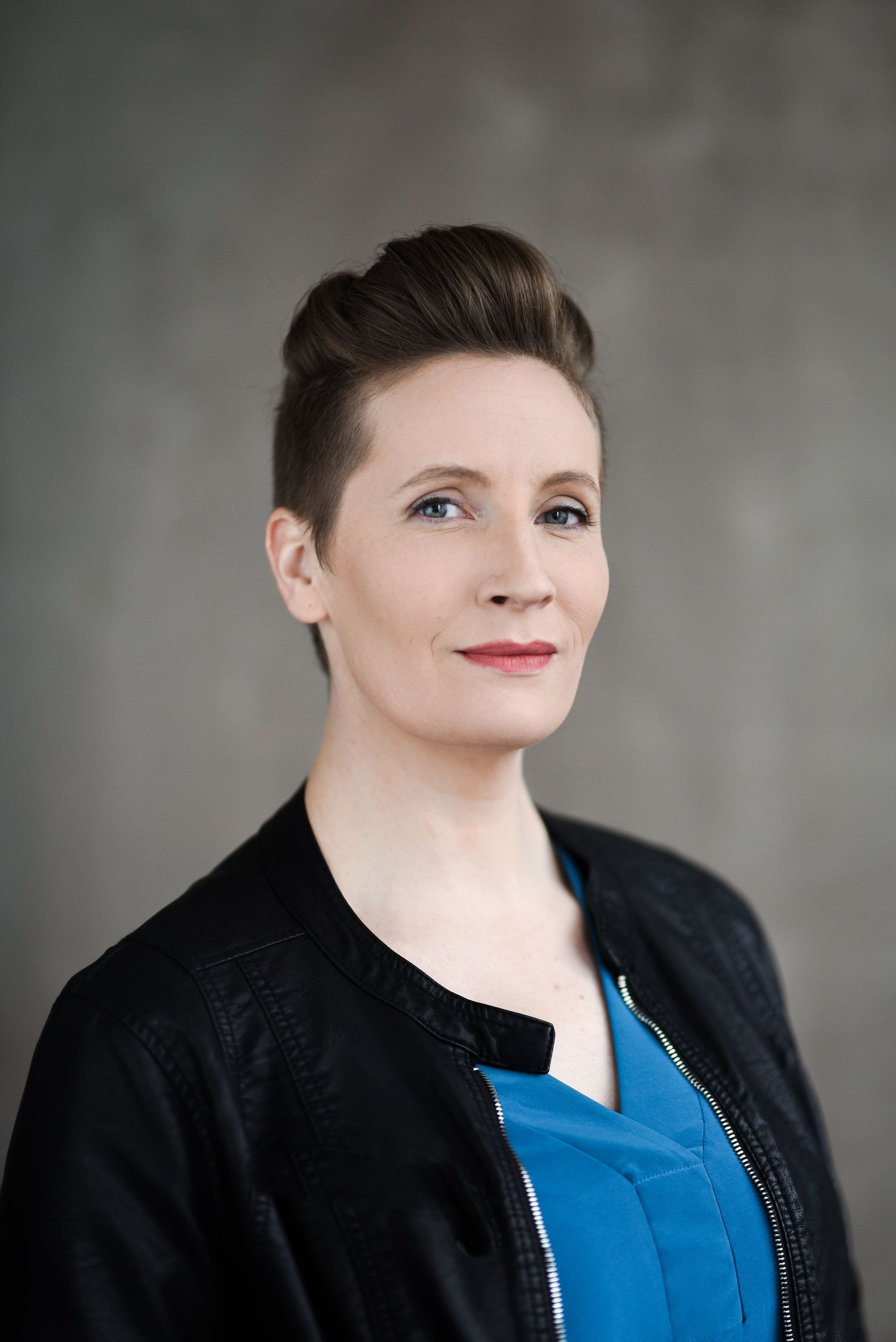 Professional headshot of person wearing black jacket and blue top with short styled hair.