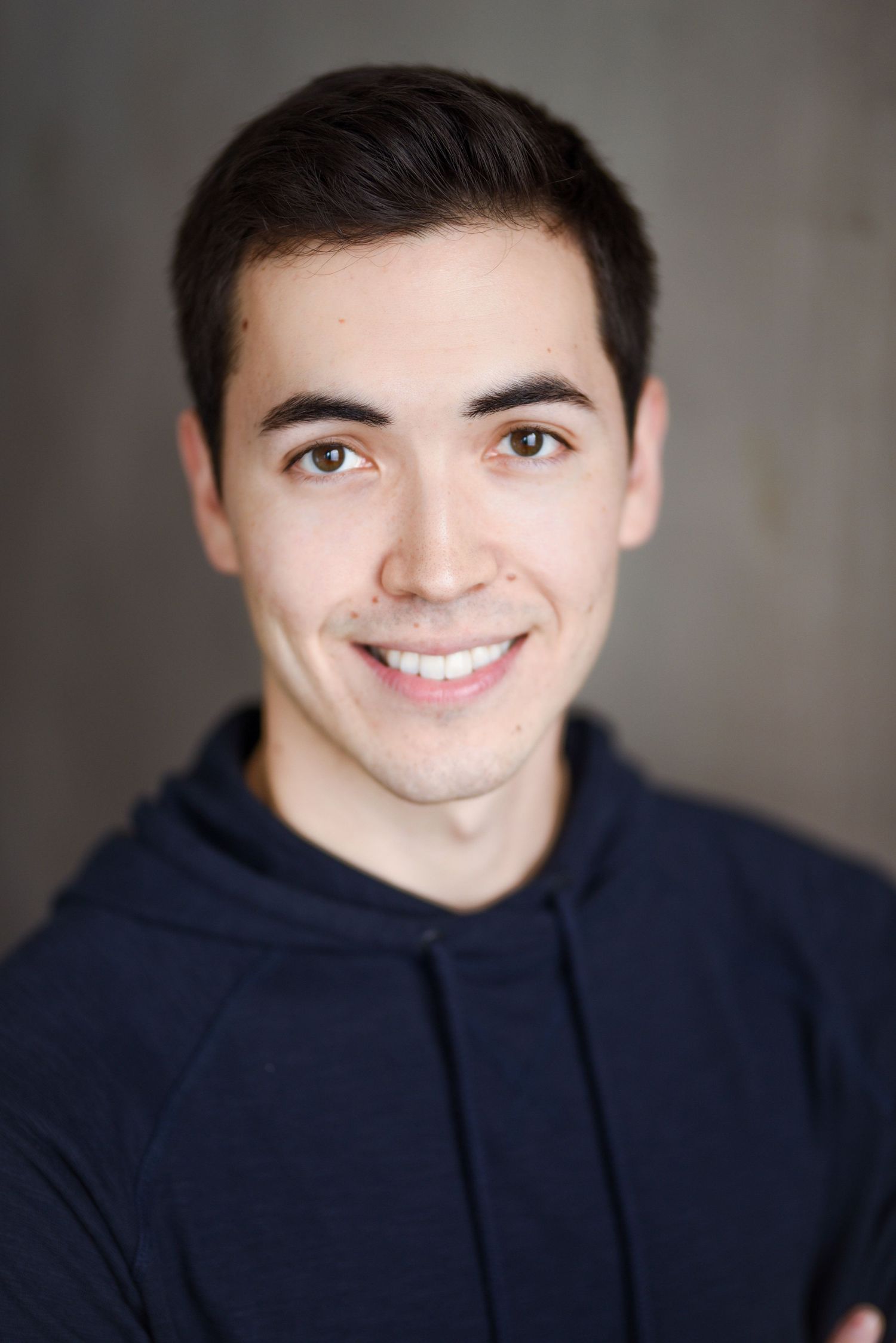 Professional portrait headshot of someone with dark hair wearing a navy blue hoodie against a grey background and smiling warmly.
