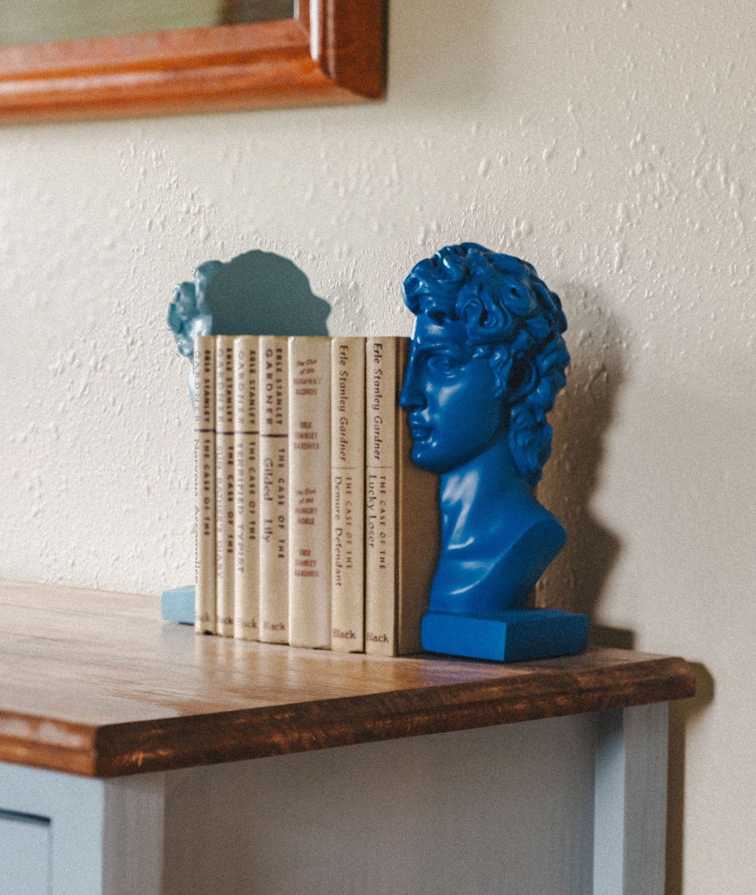 Blue decorative bust bookends holding a collection of matching books on wooden shelf.