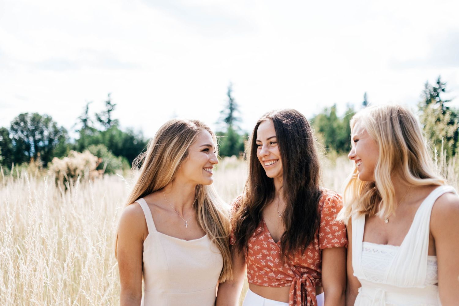 Three friends laugh together while standing in a sunlit field of tall grass on a summer day.