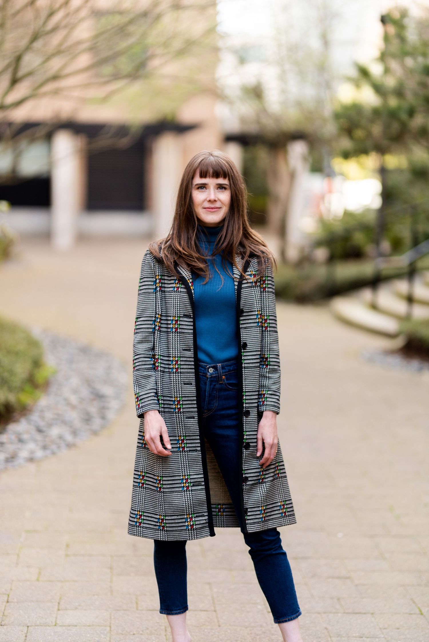 A person wearing a plaid long coat over a blue turtleneck and jeans stands on a garden path.