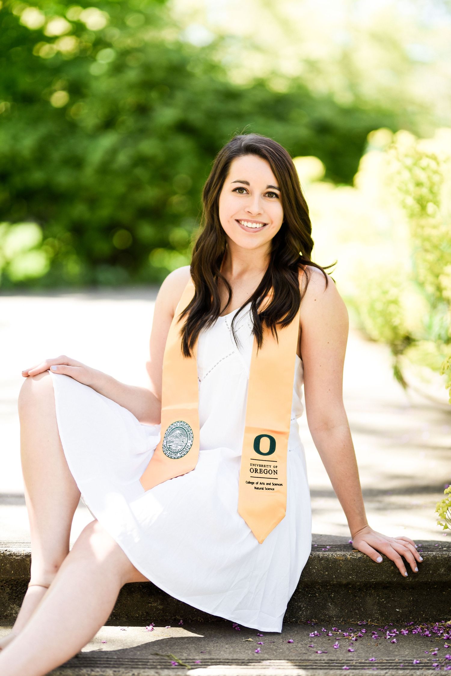 Graduate in white dress with Oregon sash sitting on stone wall.