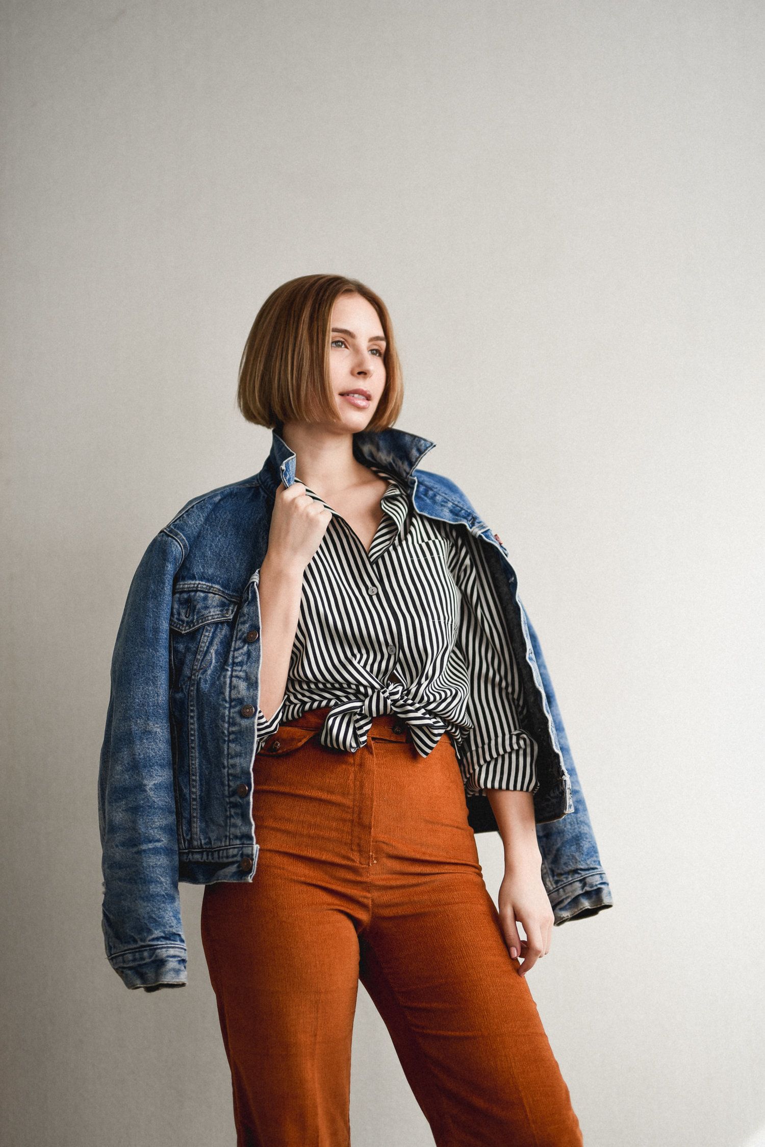 Fashion model wearing striped shirt, orange pants and denim jacket in minimalist studio setting.