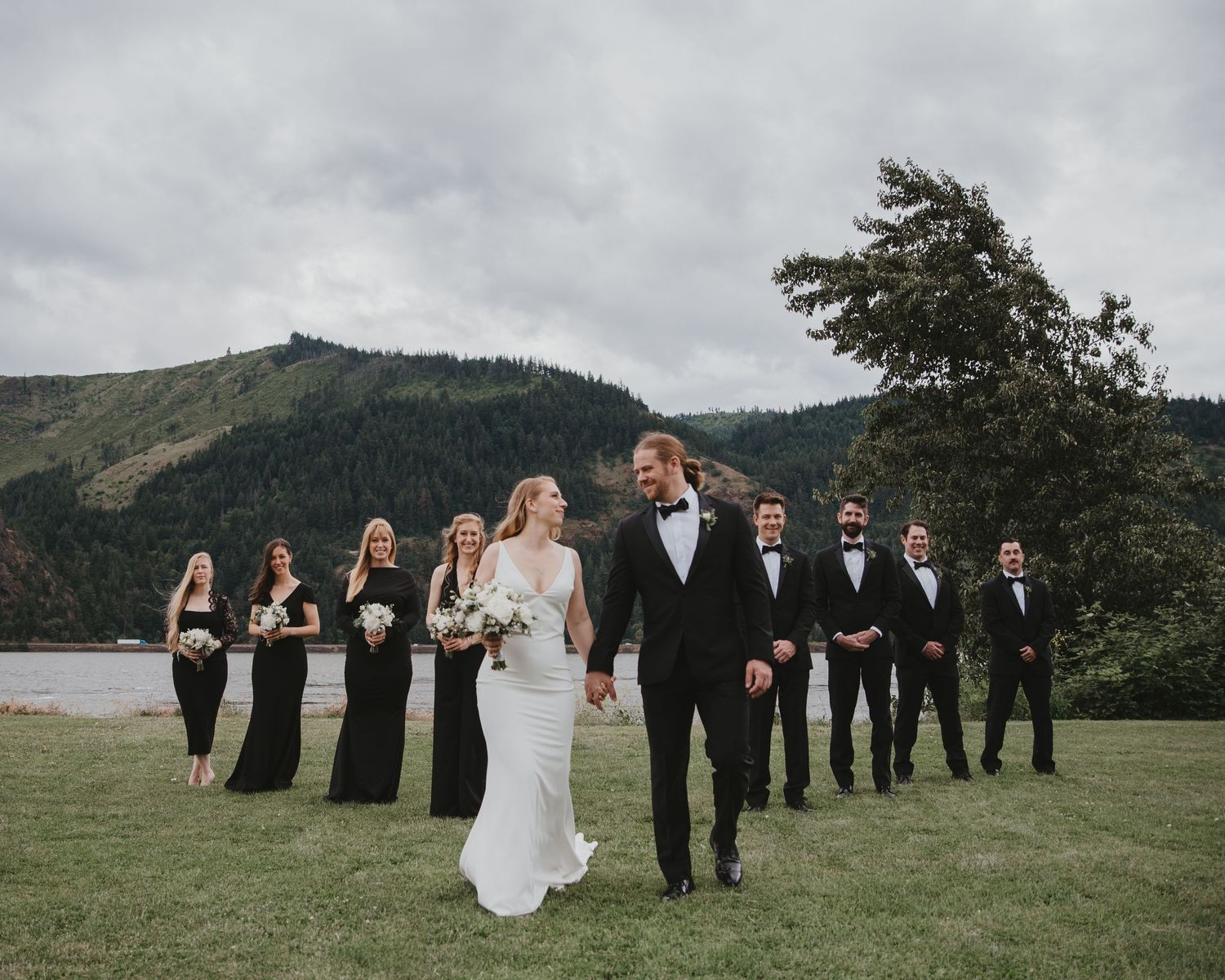 A wedding party walks across a grassy field with mountains and a lake in the background.