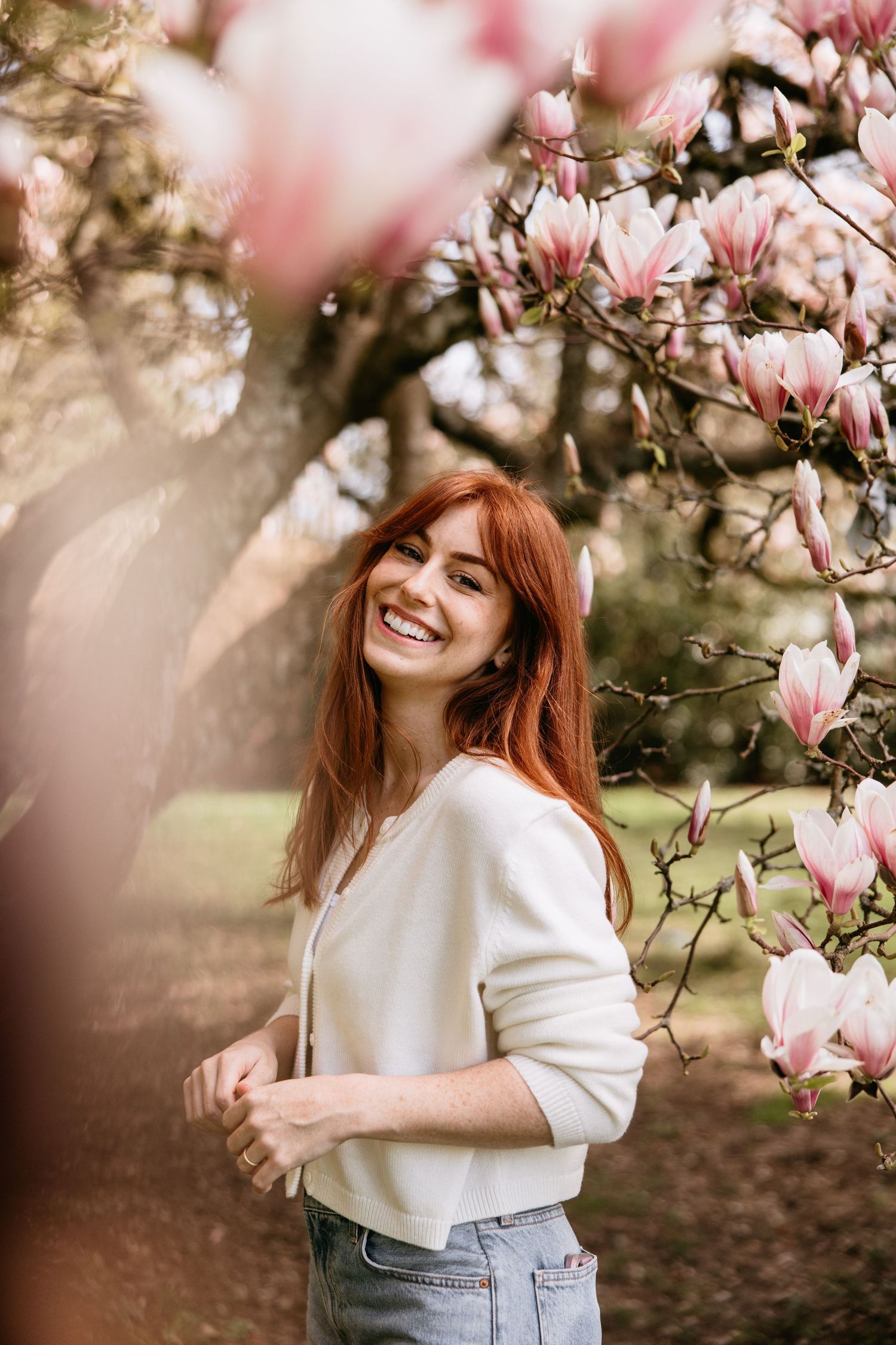 A woman in a white sweater poses joyfully near blooming pink magnolia flowers in springtime.