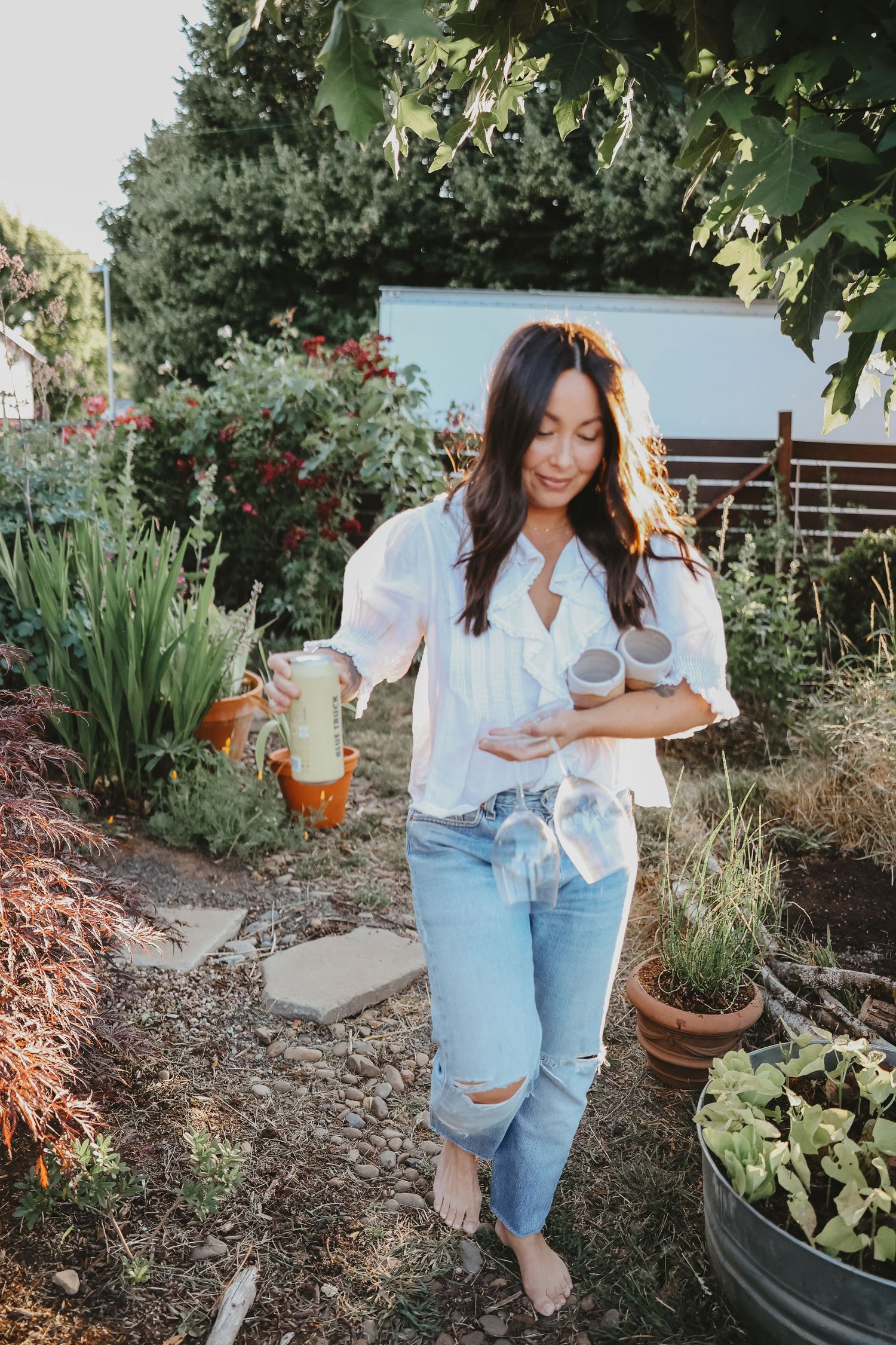 Person in white shirt and ripped jeans holding drinks in a lush garden setting.