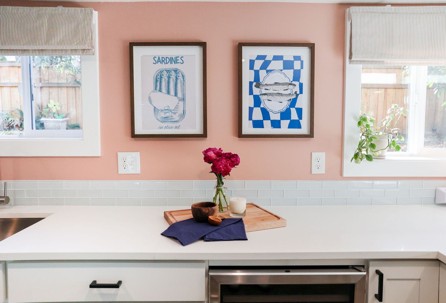 Modern kitchen with white counters, pink walls, and blue framed artwork displaying dishes and cookware.