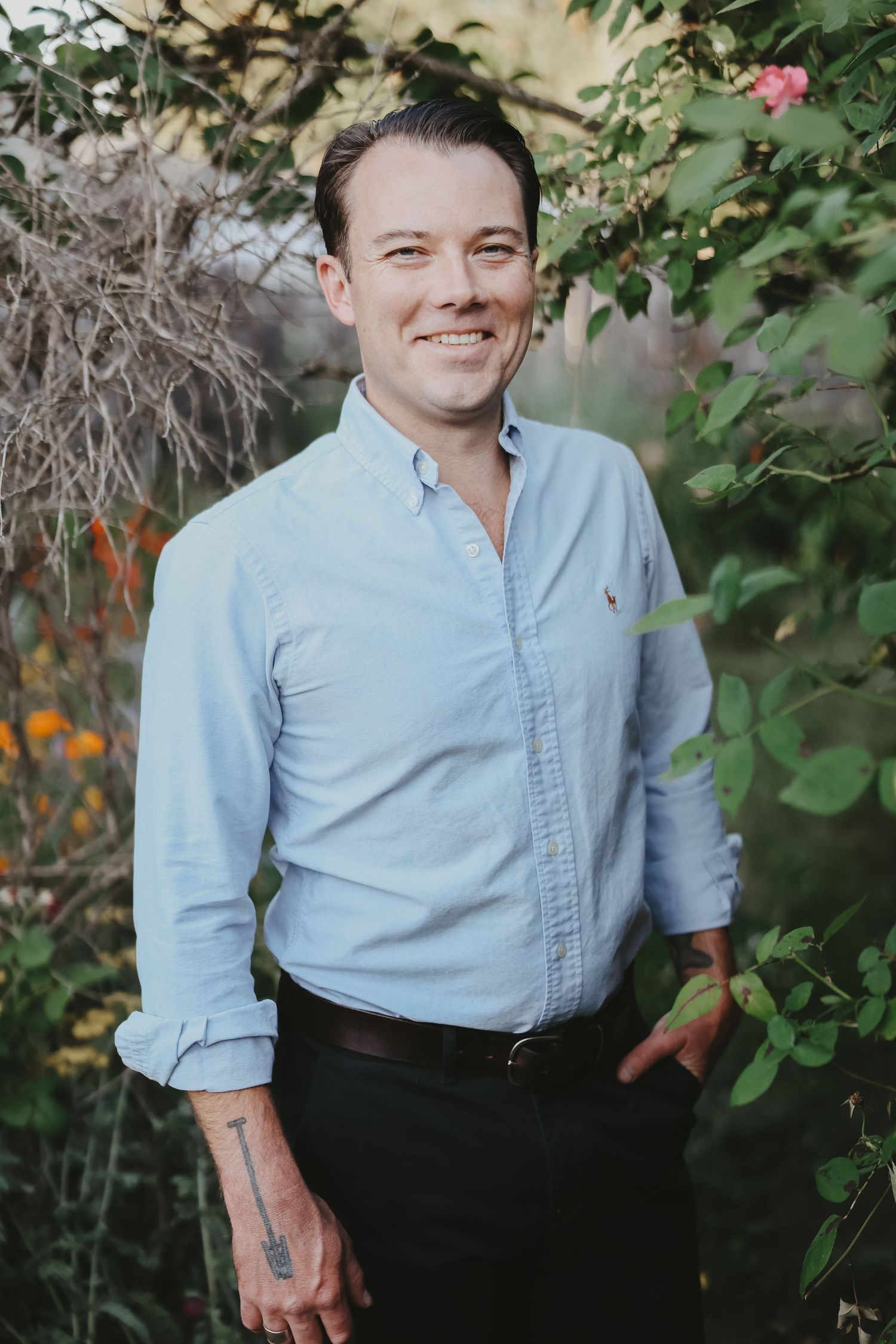Person in light blue button-down shirt smiling in garden setting with flowers.