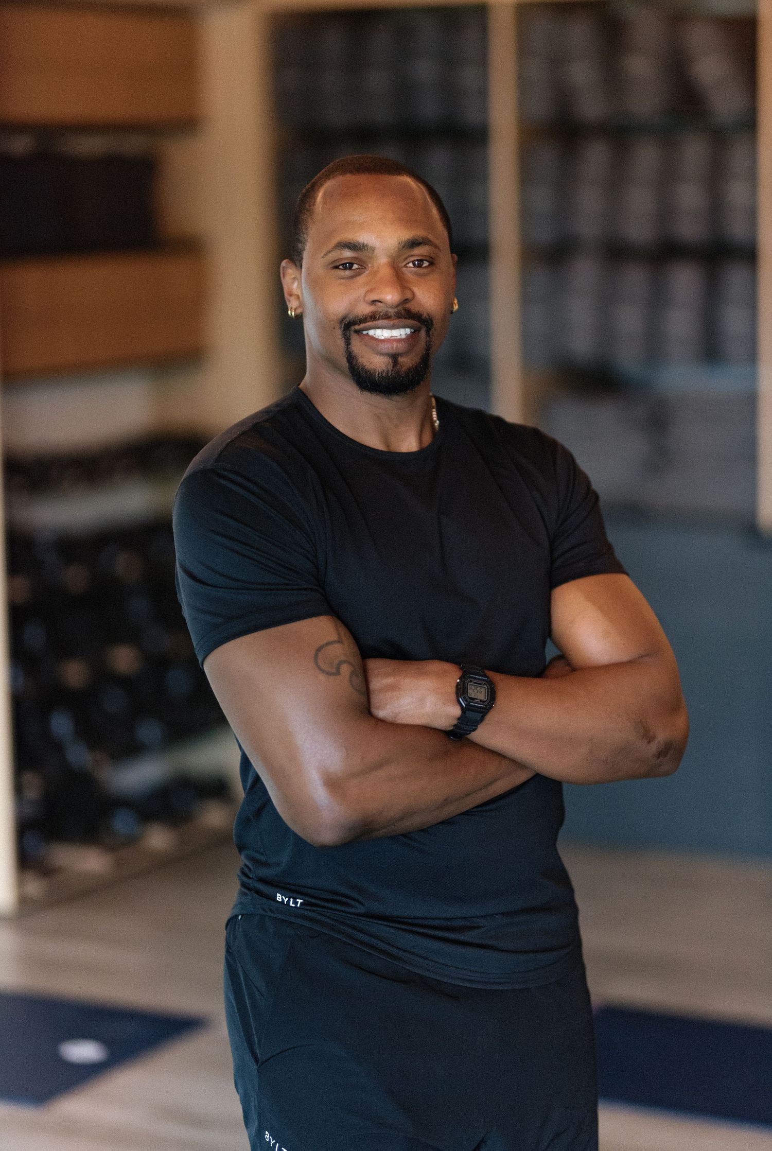 Smiling muscular man in black t-shirt stands with arms crossed in a modern gym with dumbbells in background.