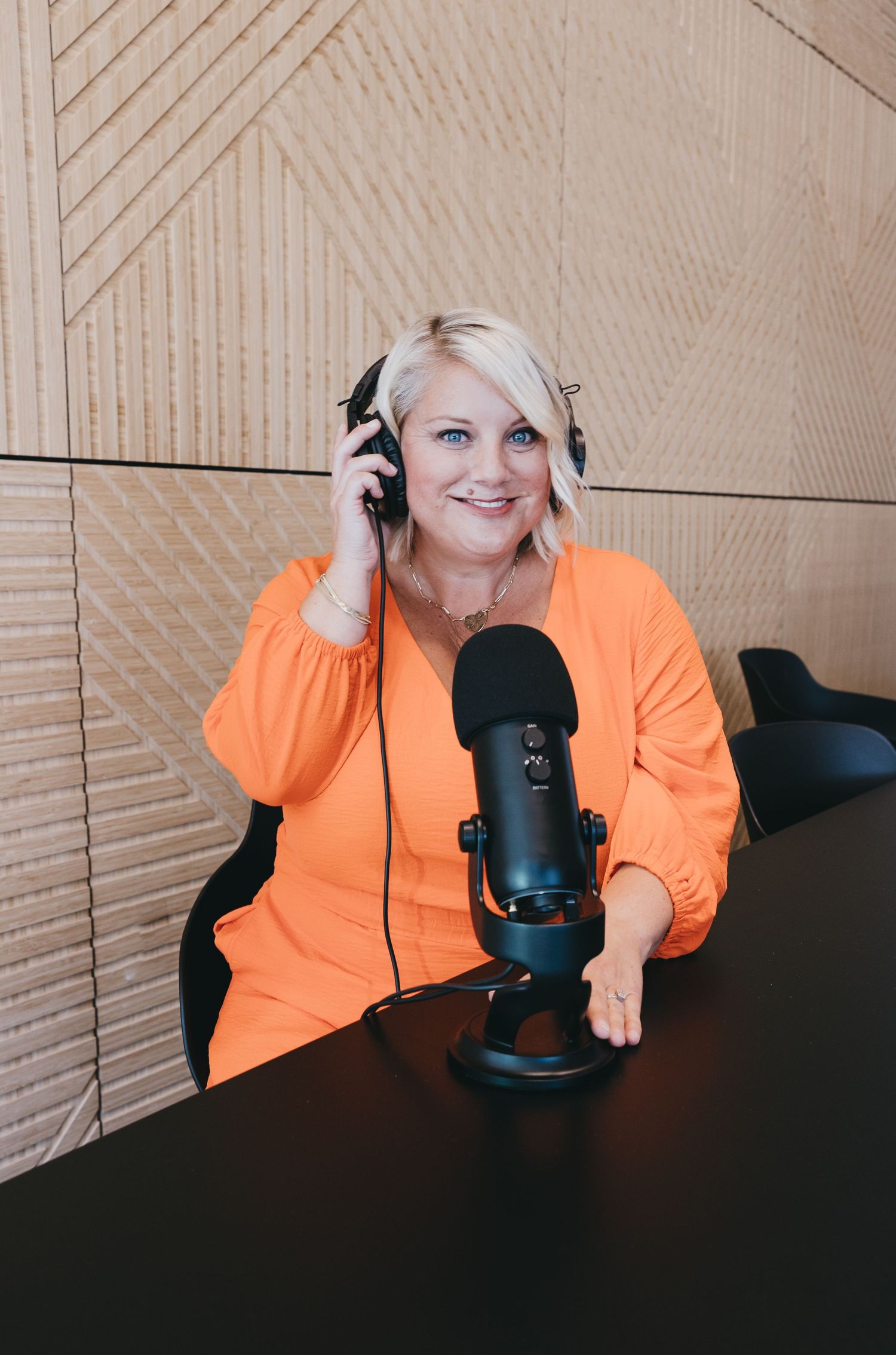 Person in bright orange sweater sits at desk with microphone and headphones while recording podcast content.