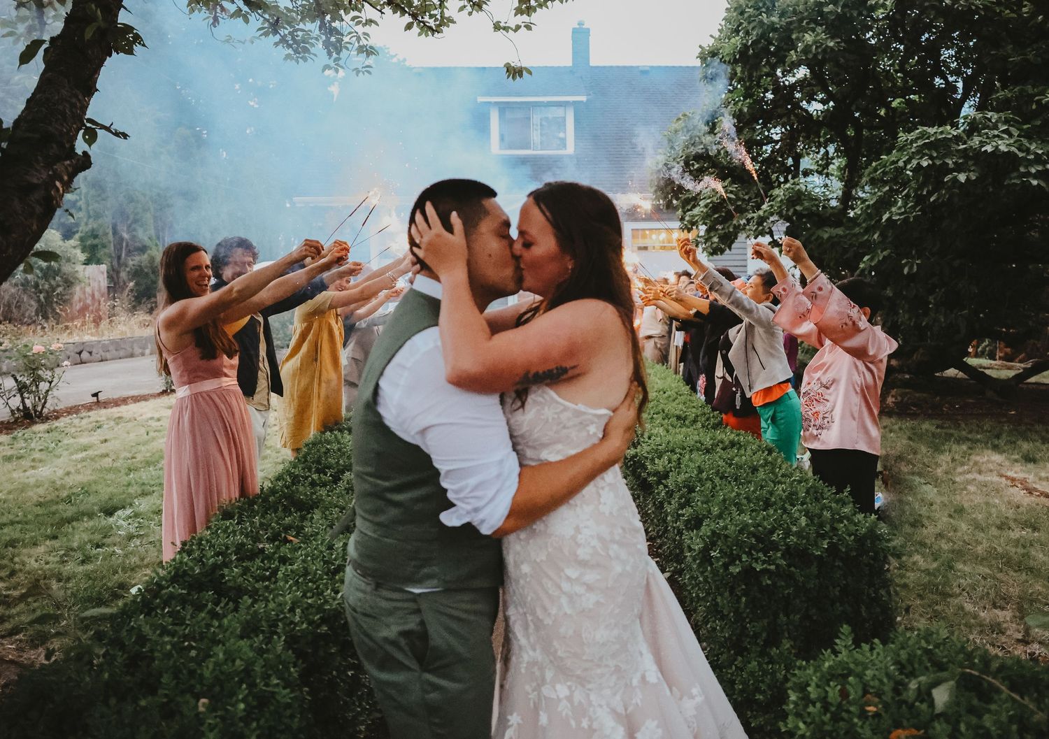 Newlyweds share a kiss while wedding guests celebrate with sparklers in an outdoor garden setting.