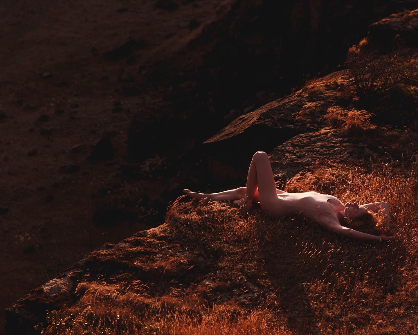 An artistic nude figure reclines on reddish-brown rocks in dramatic lighting.