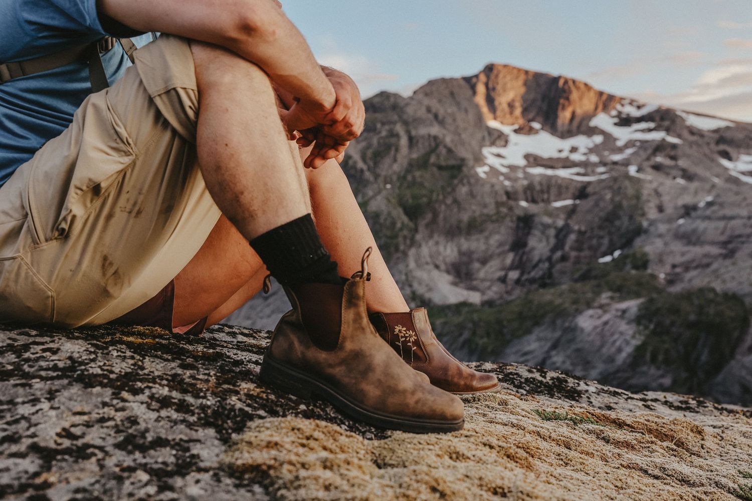 Close up of hiking boots on rocky mountain terrain with snow-capped peaks in the distance.