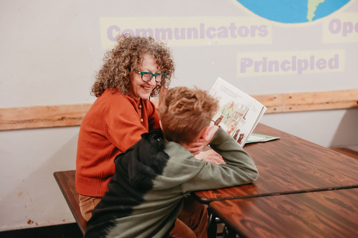 Two students share a book together at a wooden table in a classroom setting.