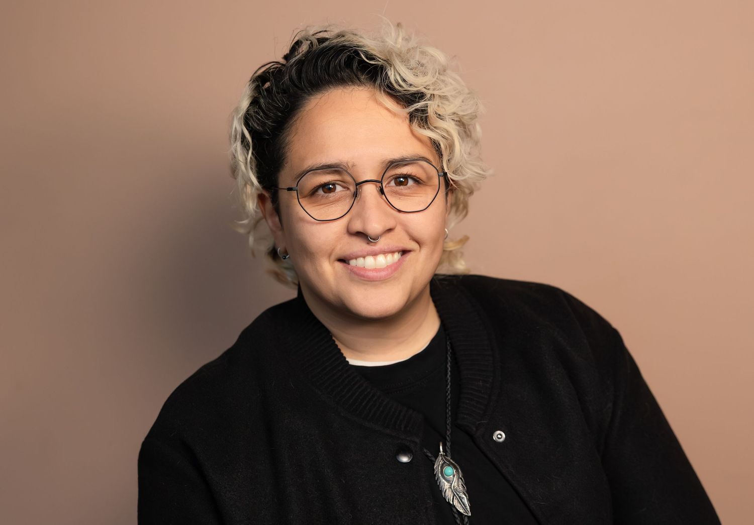 Smiling person with curly hair and glasses wearing a black jacket and teal pendant necklace against a beige background.
