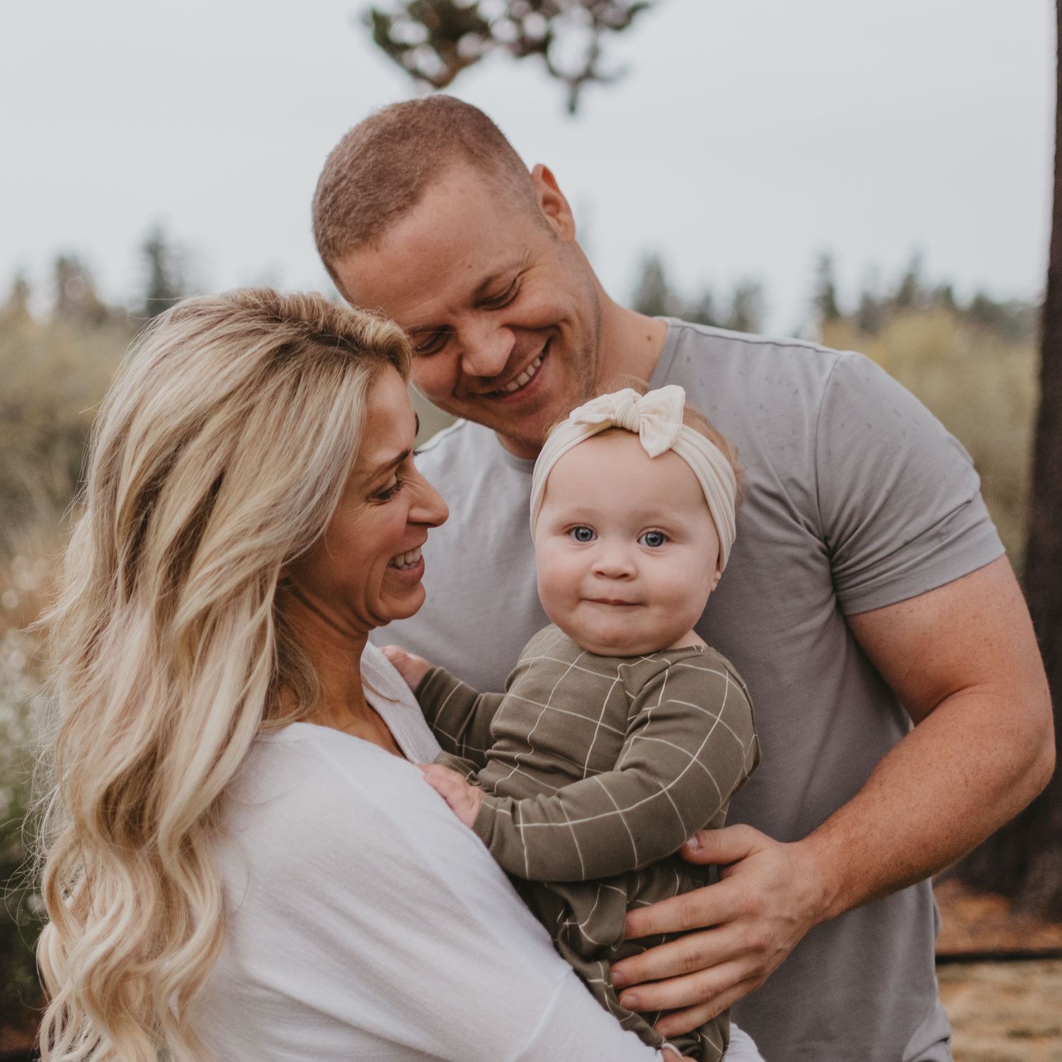 Parents share an intimate outdoor moment while holding their smiling baby in casual attire.