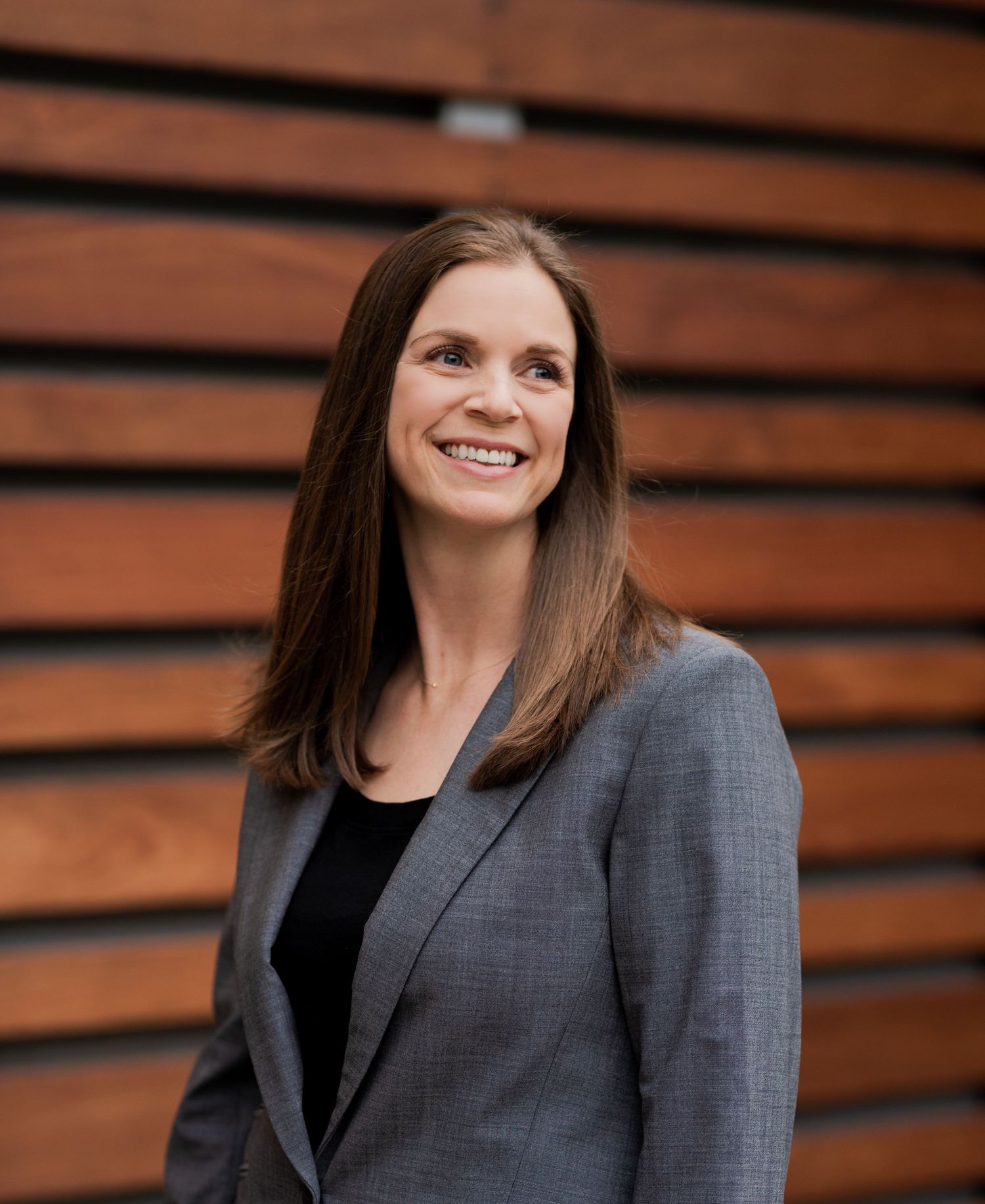 Professional headshot of person in grey blazer smiling against wooden wall background.