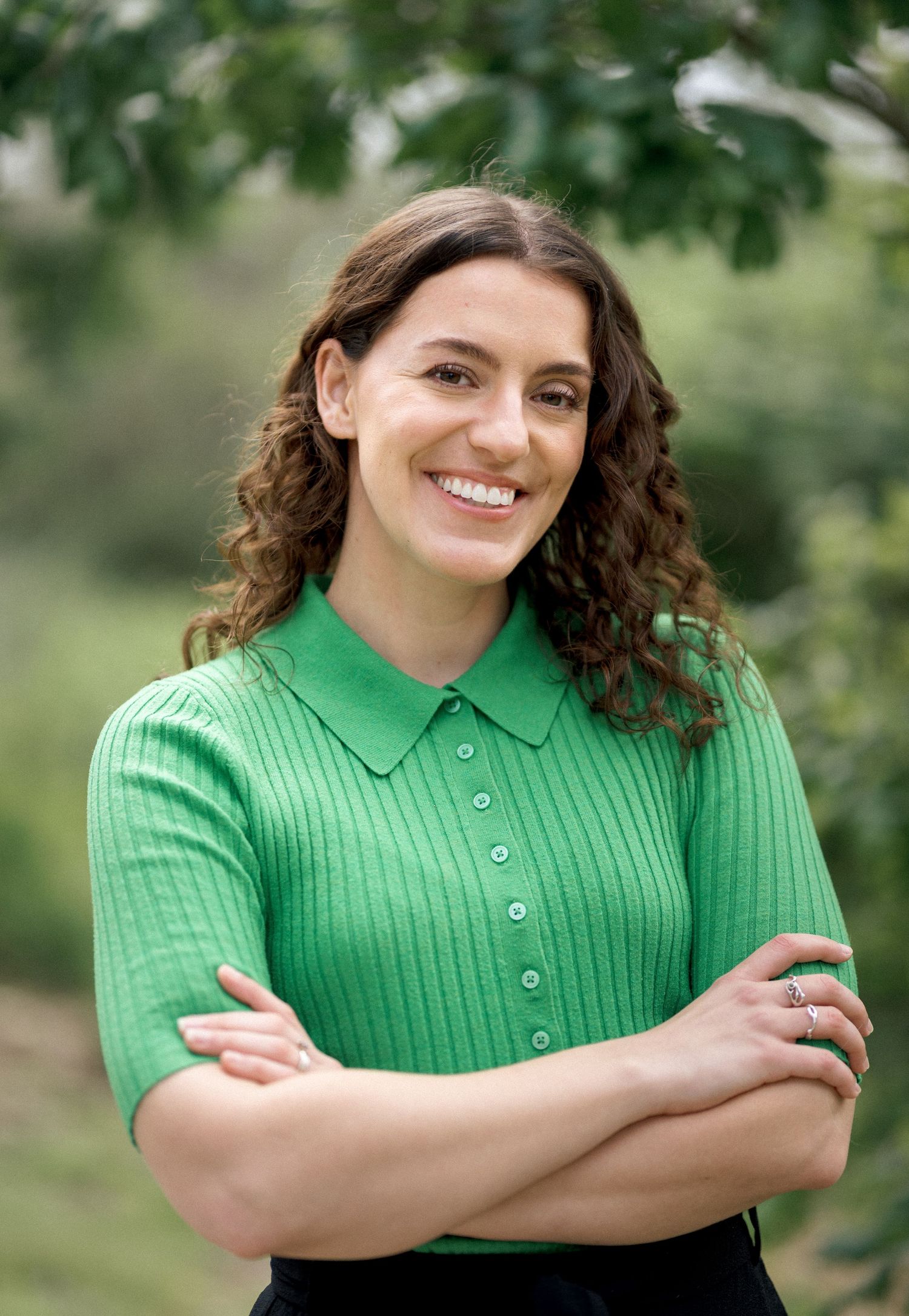 Smiling woman with curly hair in green polo shirt, arms crossed, posing outdoors with lush green foliage background.