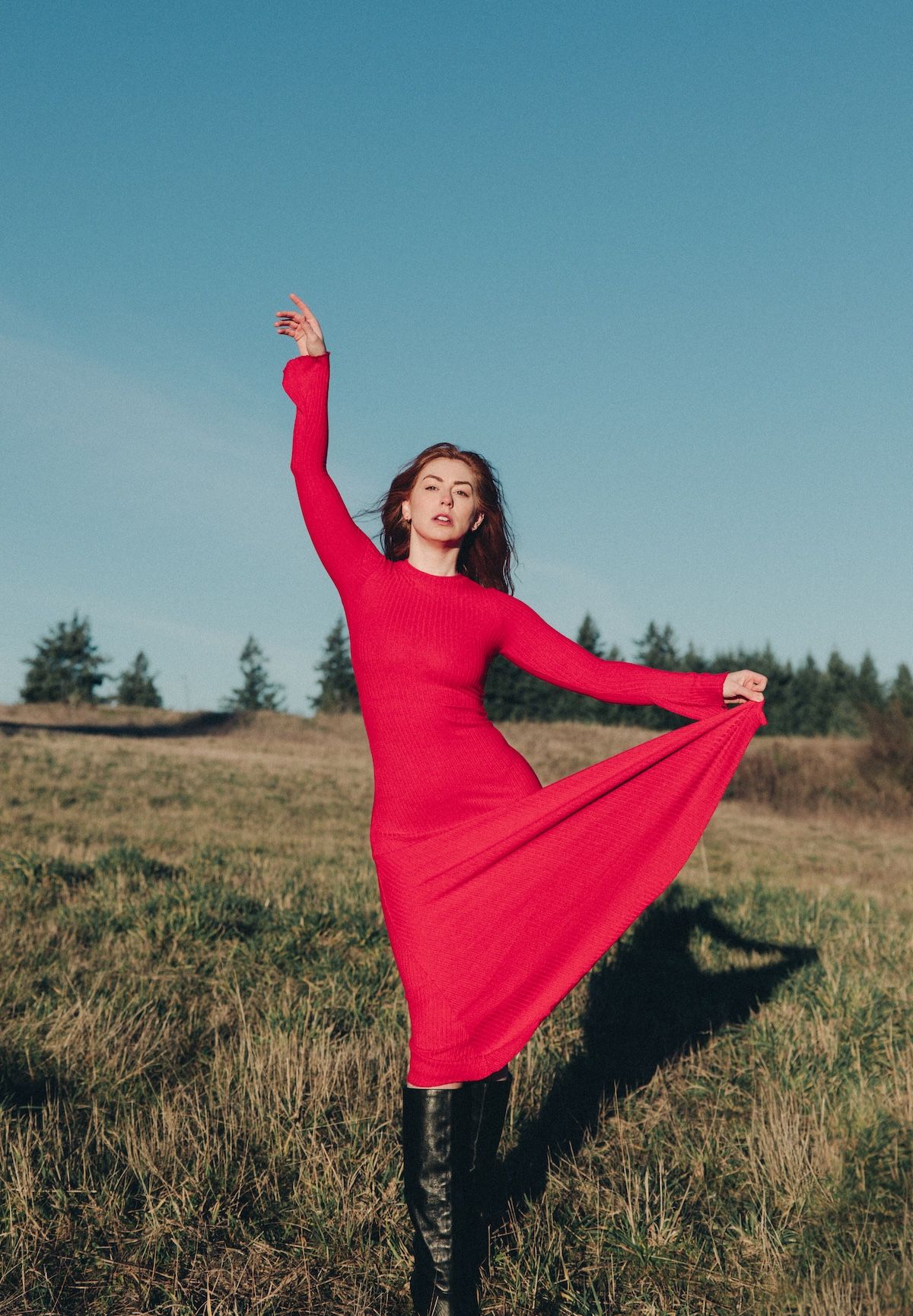 A figure in a flowing red dress twirls gracefully in a grassy meadow against a blue sky background.