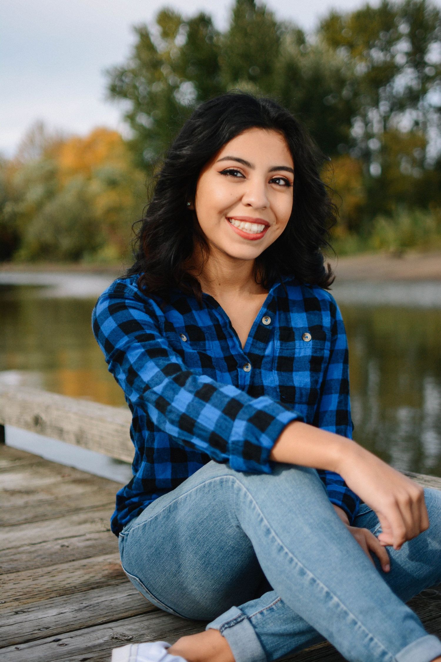 Casual portrait in blue plaid shirt and jeans sitting on wooden dock by autumn lake.