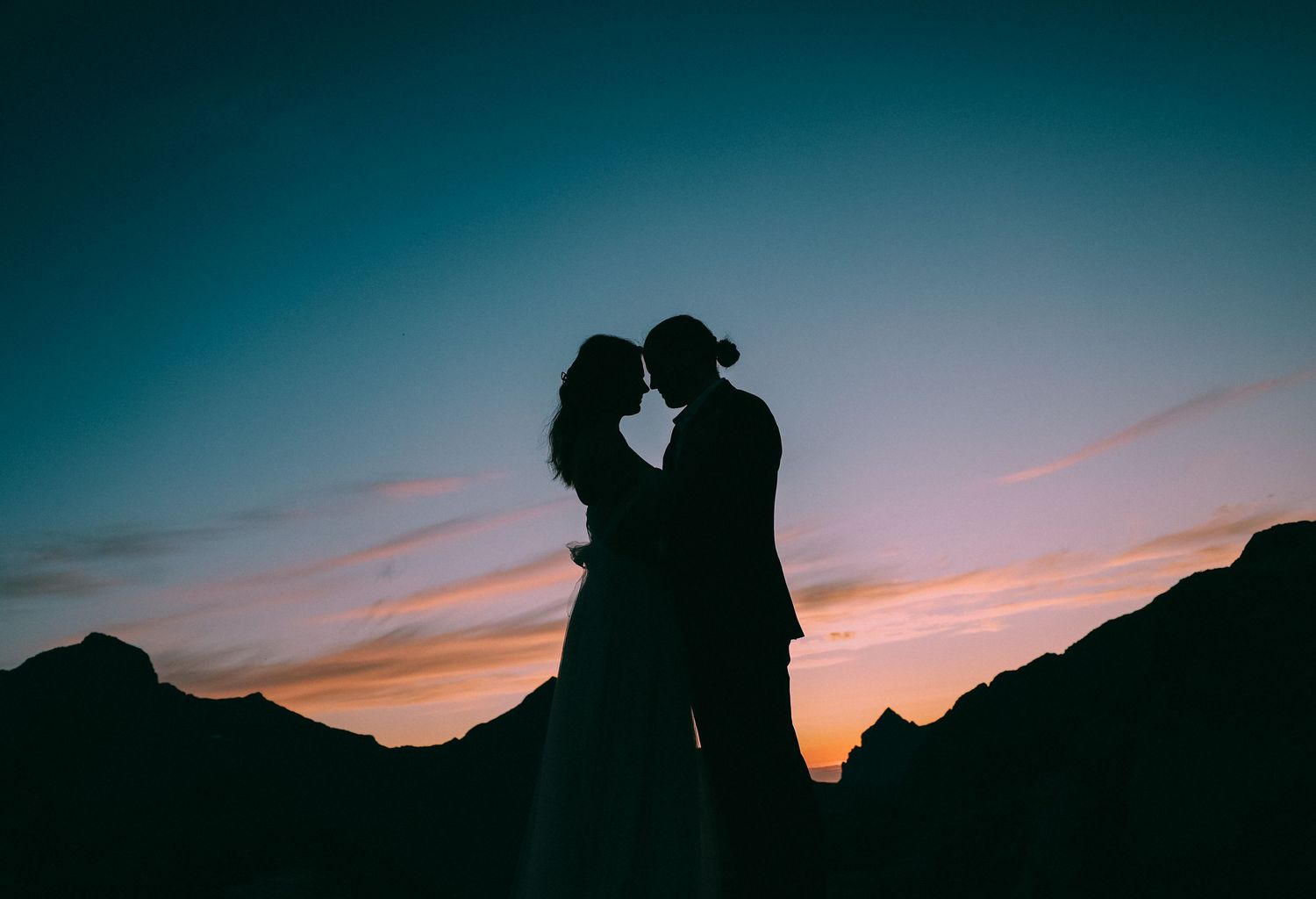 Silhouette of a couple against a vibrant sunset sky with mountain peaks in the background.