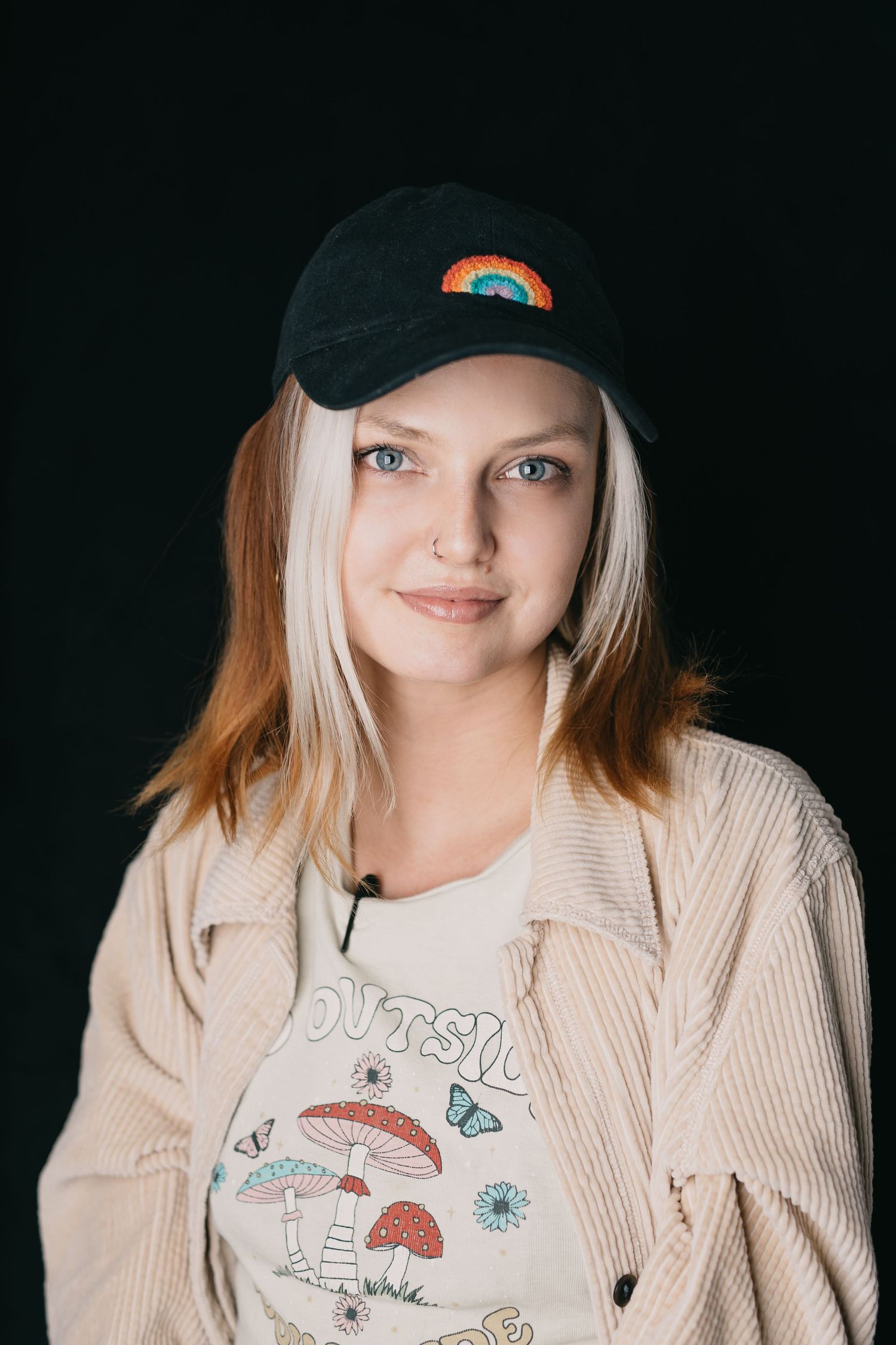 Studio portrait wearing a black baseball cap with rainbow design and beige cardigan.