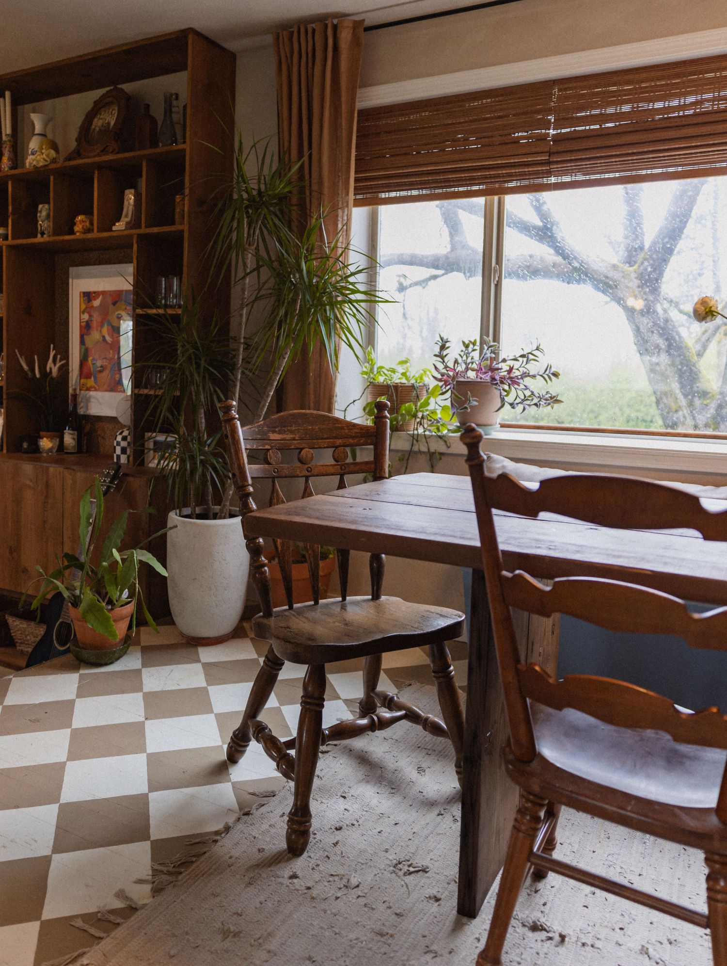 Rustic dining area with checkerboard floor and wooden chairs near a large window.