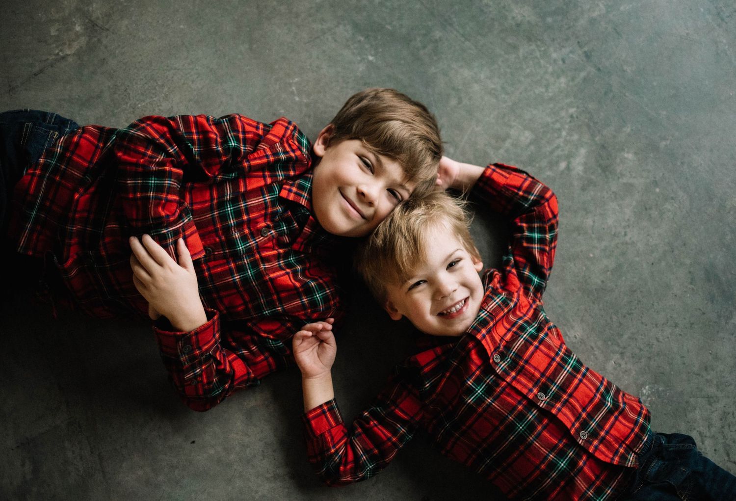 Two children in matching red plaid outfits lying on gray floor.