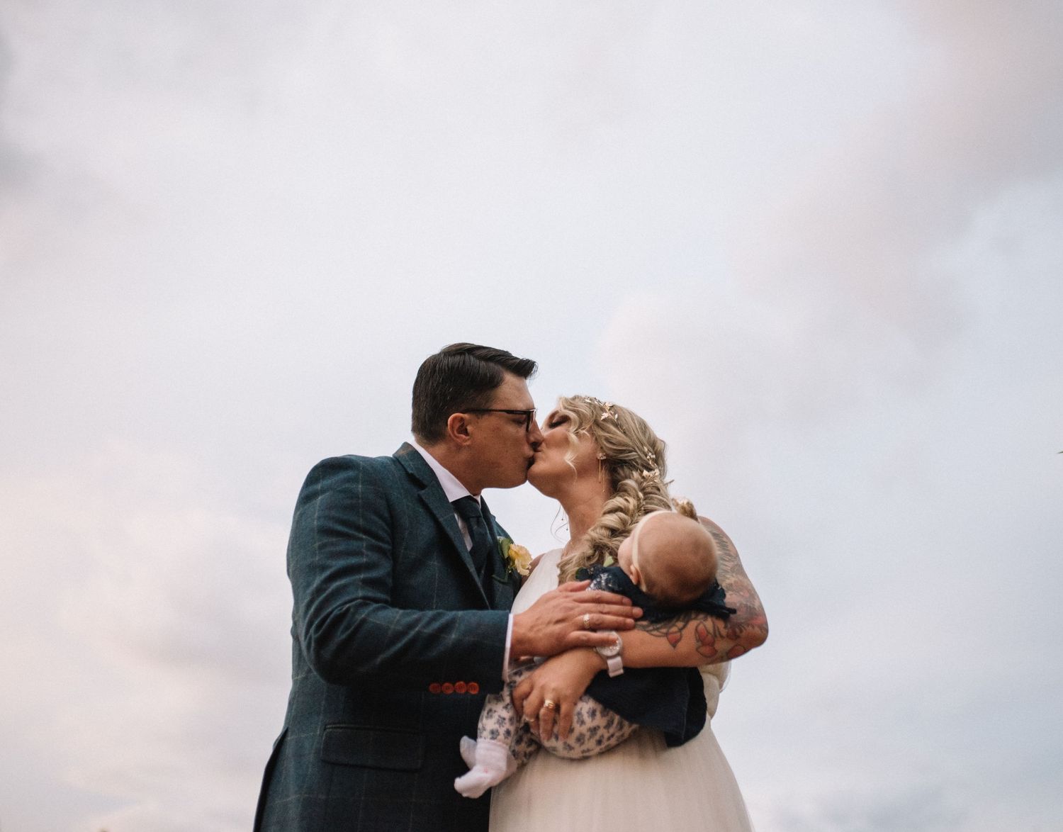A couple in wedding attire share a romantic kiss against a pink sunset sky.