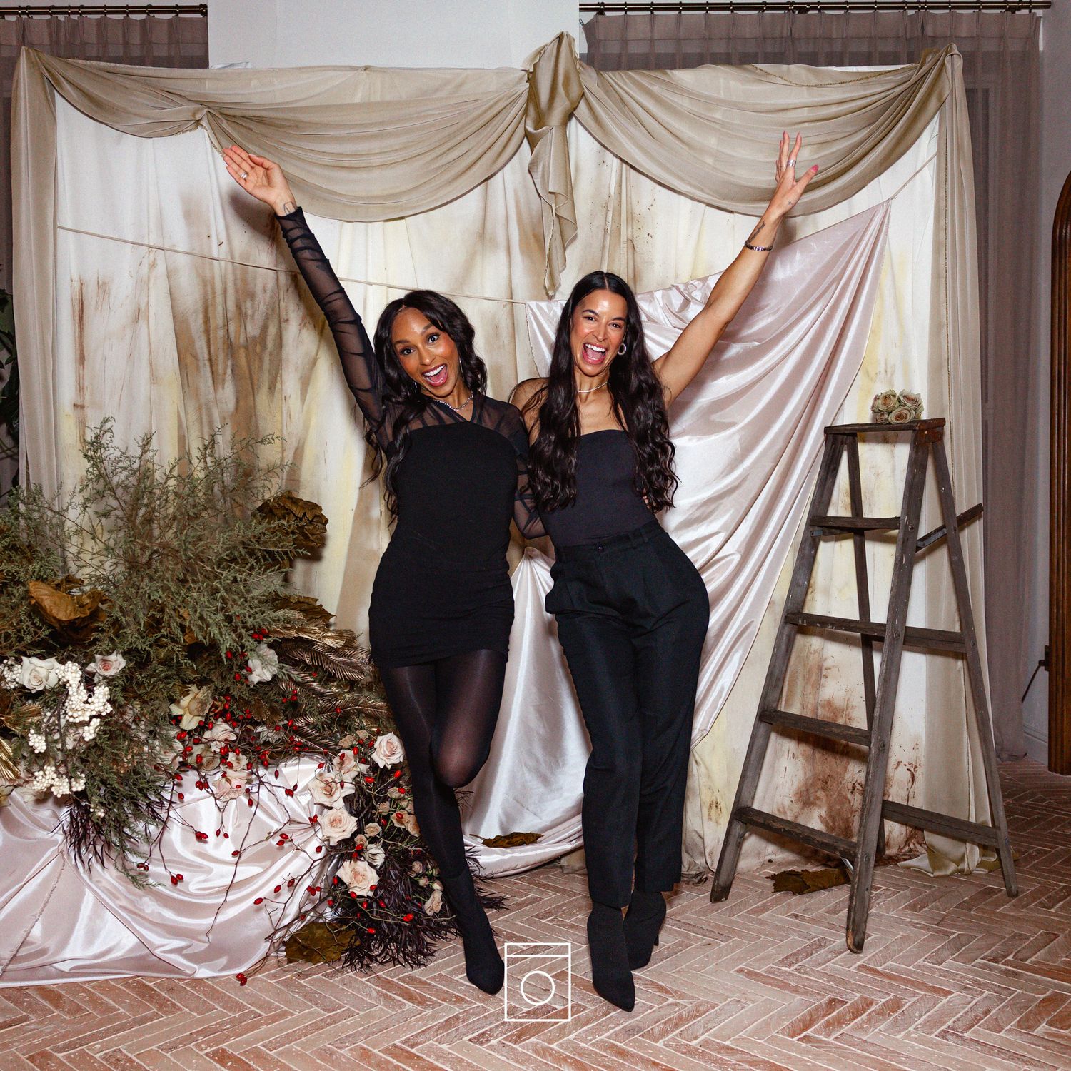 Two people in black outfits pose playfully in front of white draped fabric and wooden ladders in a studio setting.