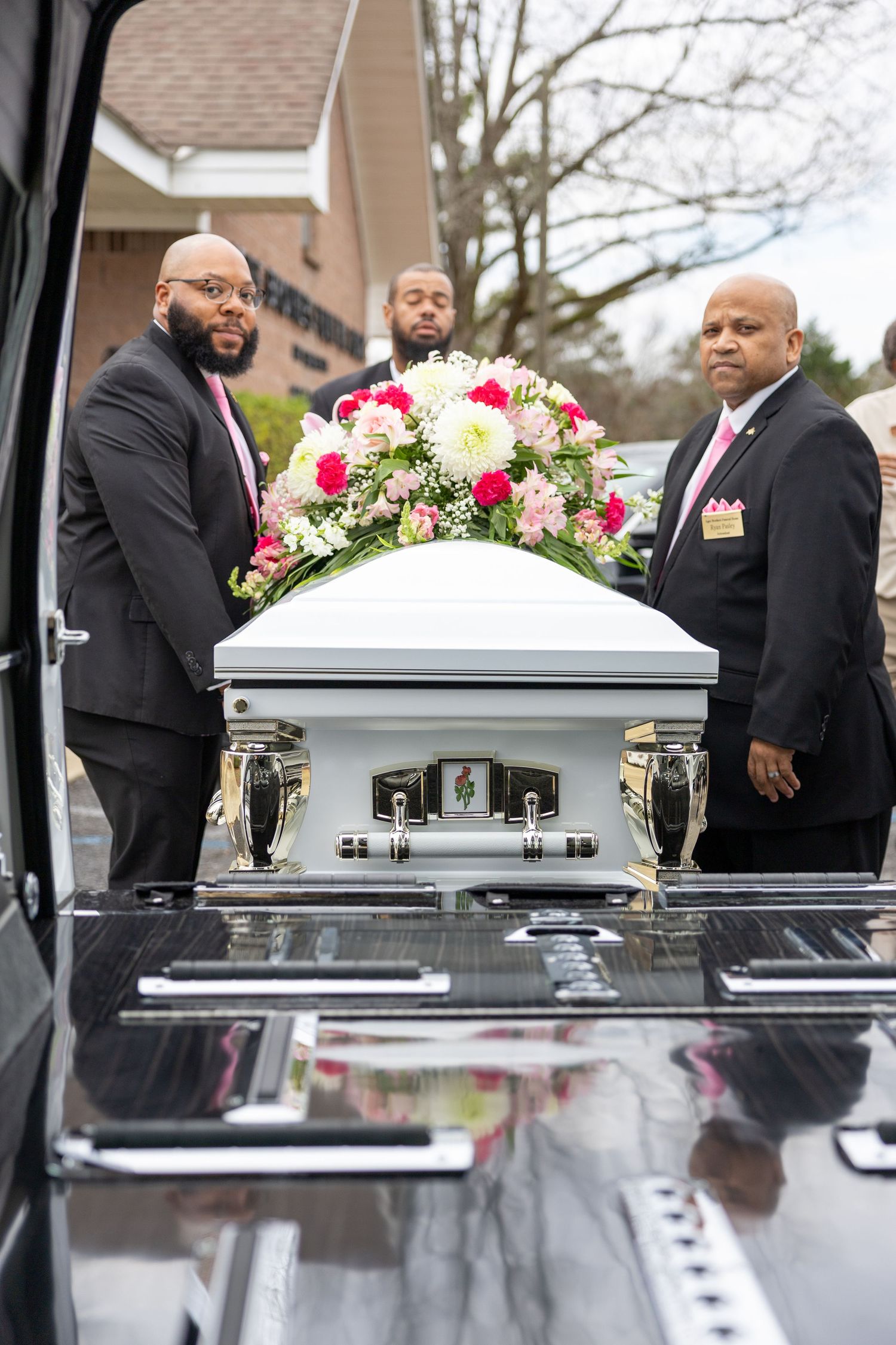 White casket adorned with pink and white floral arrangements being carried by pallbearers at a funeral service.