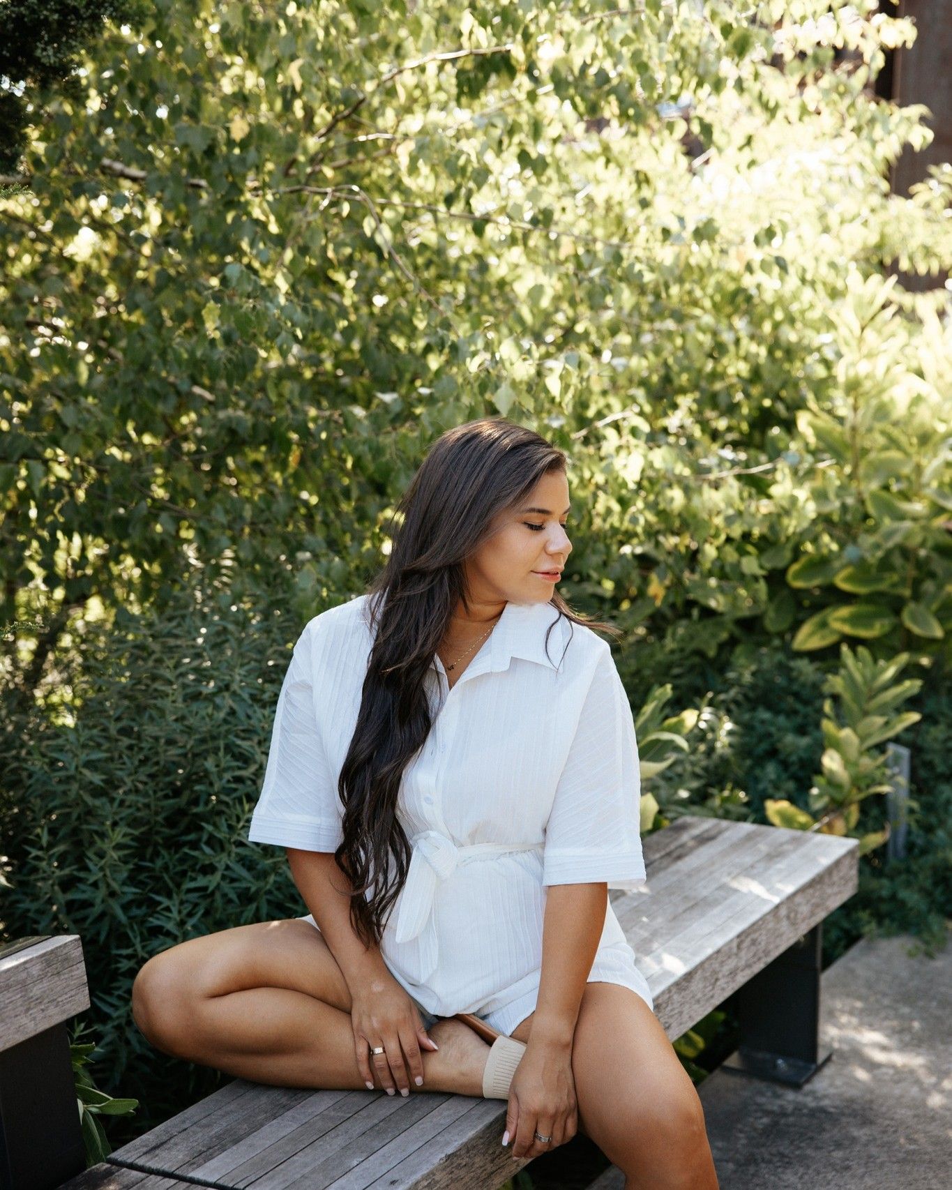 Woman in white shirt romper sitting on wooden bench surrounded by lush green foliage in natural light.