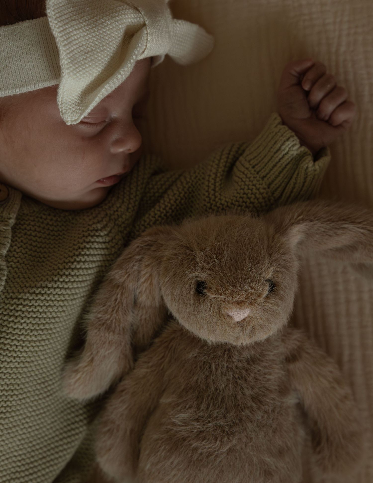 Sleeping newborn baby in green knit sweater and bow headband cuddled next to a soft brown bunny stuffed animal.