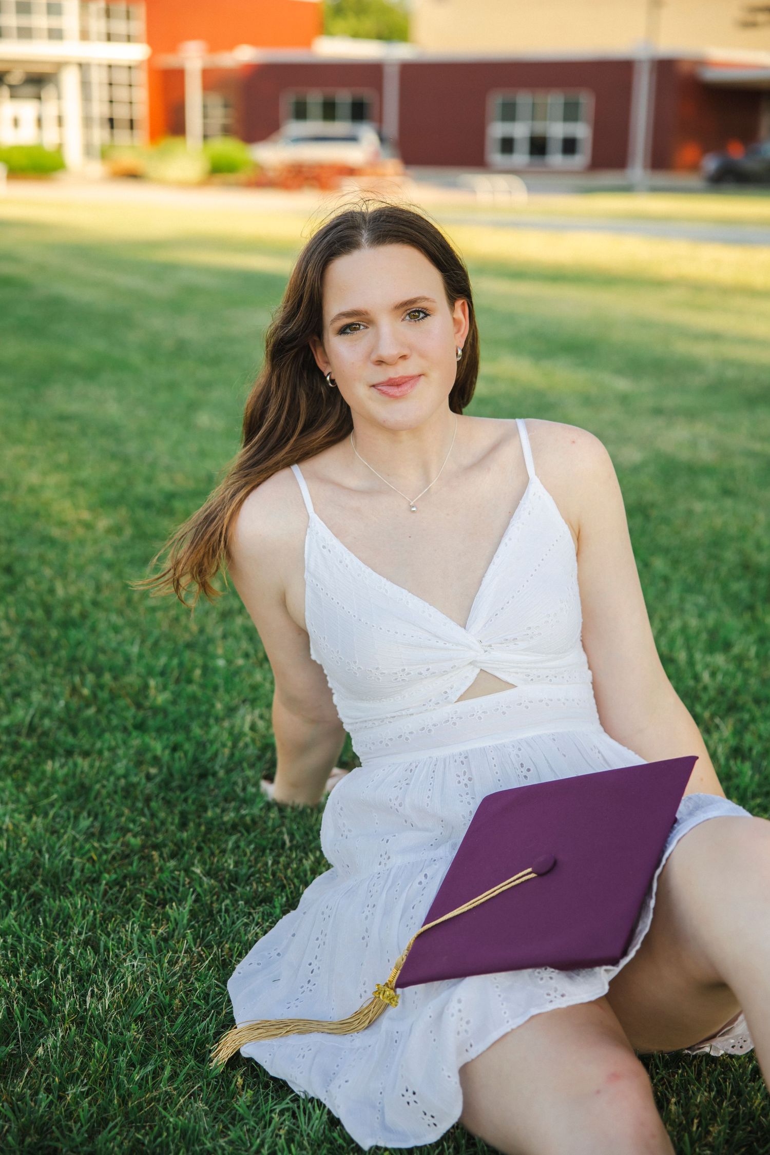 Graduate in white eyelet dress sits on grass holding purple diploma cover during sunset photo session.