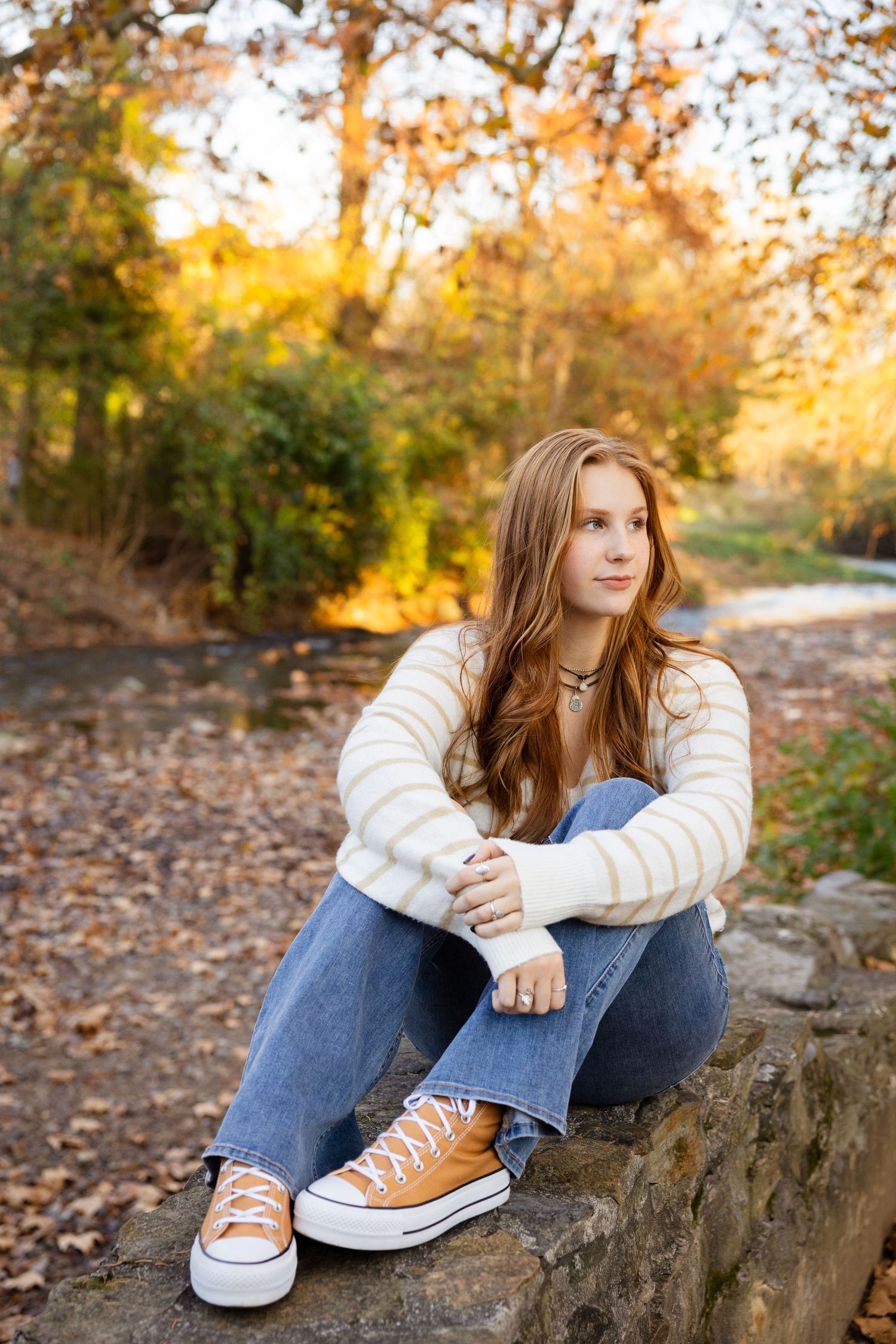 Autumn outdoor portrait sitting on rocks wearing jeans, tan sneakers and a striped sweater.