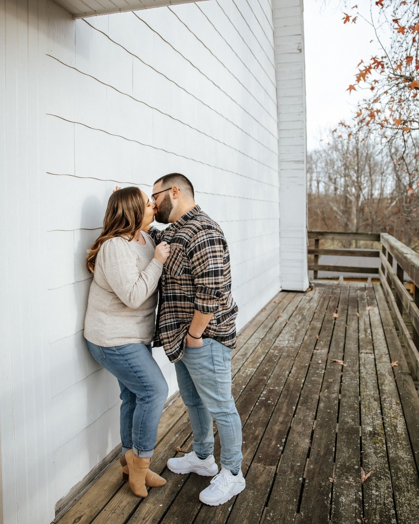 A couple shares a romantic kiss leaning against a white shiplap wall on a wooden deck in autumn.