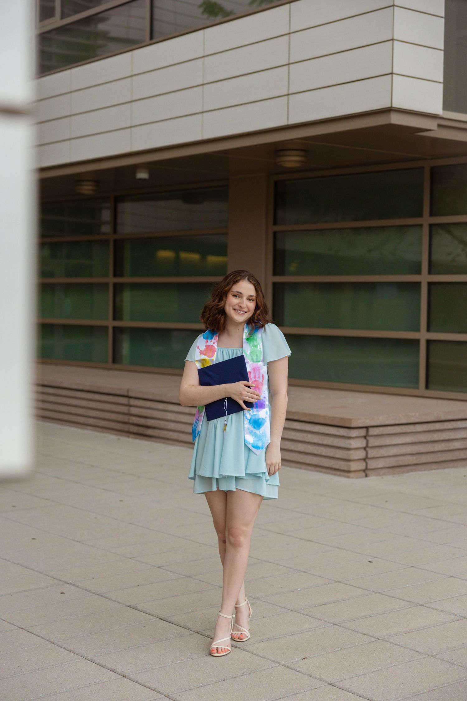 A graduate in a mint dress poses playfully outside a modern campus building.