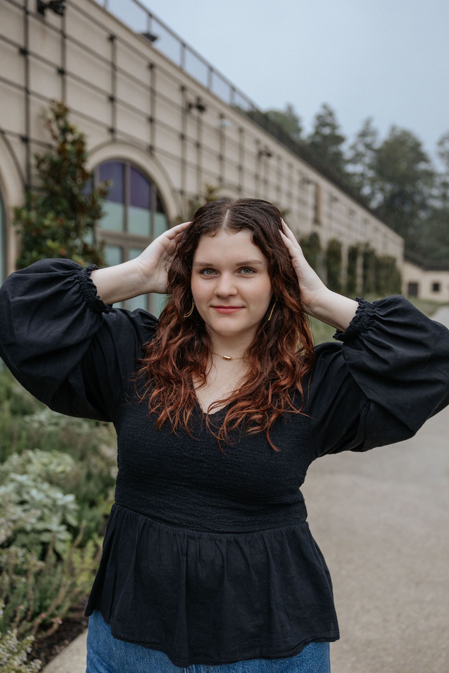 Casual outdoor portrait in black peplum top with hands in hair near garden architecture.
