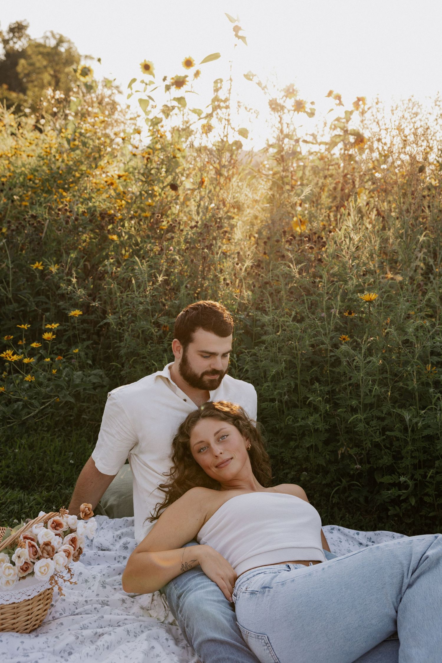 Couple relaxing in a sunflower field at golden hour, woman lying down with floral crown, man sitting beside her.