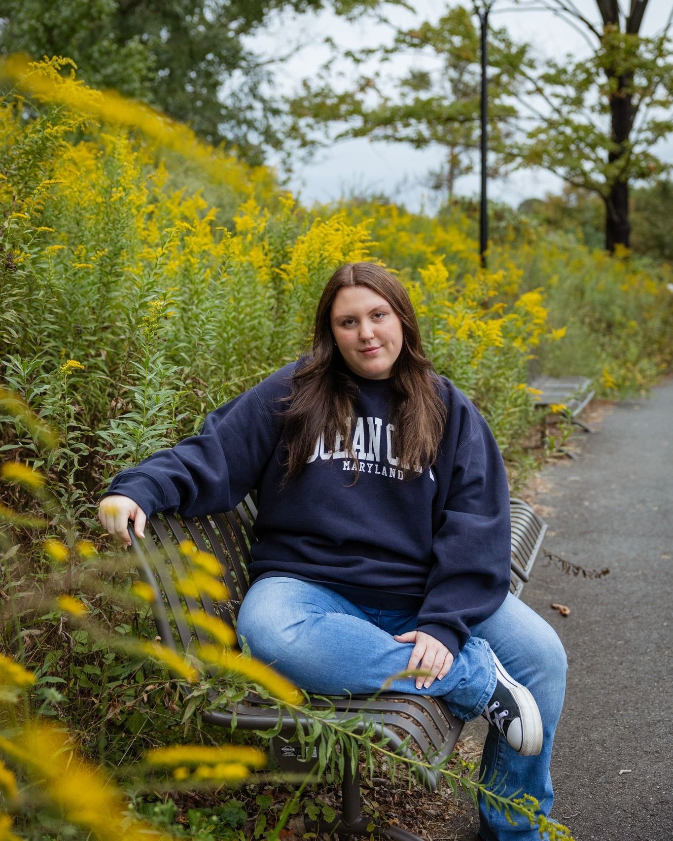 Young woman in navy University of Maryland hoodie sitting on bench surrounded by yellow wildflowers along a scenic path.