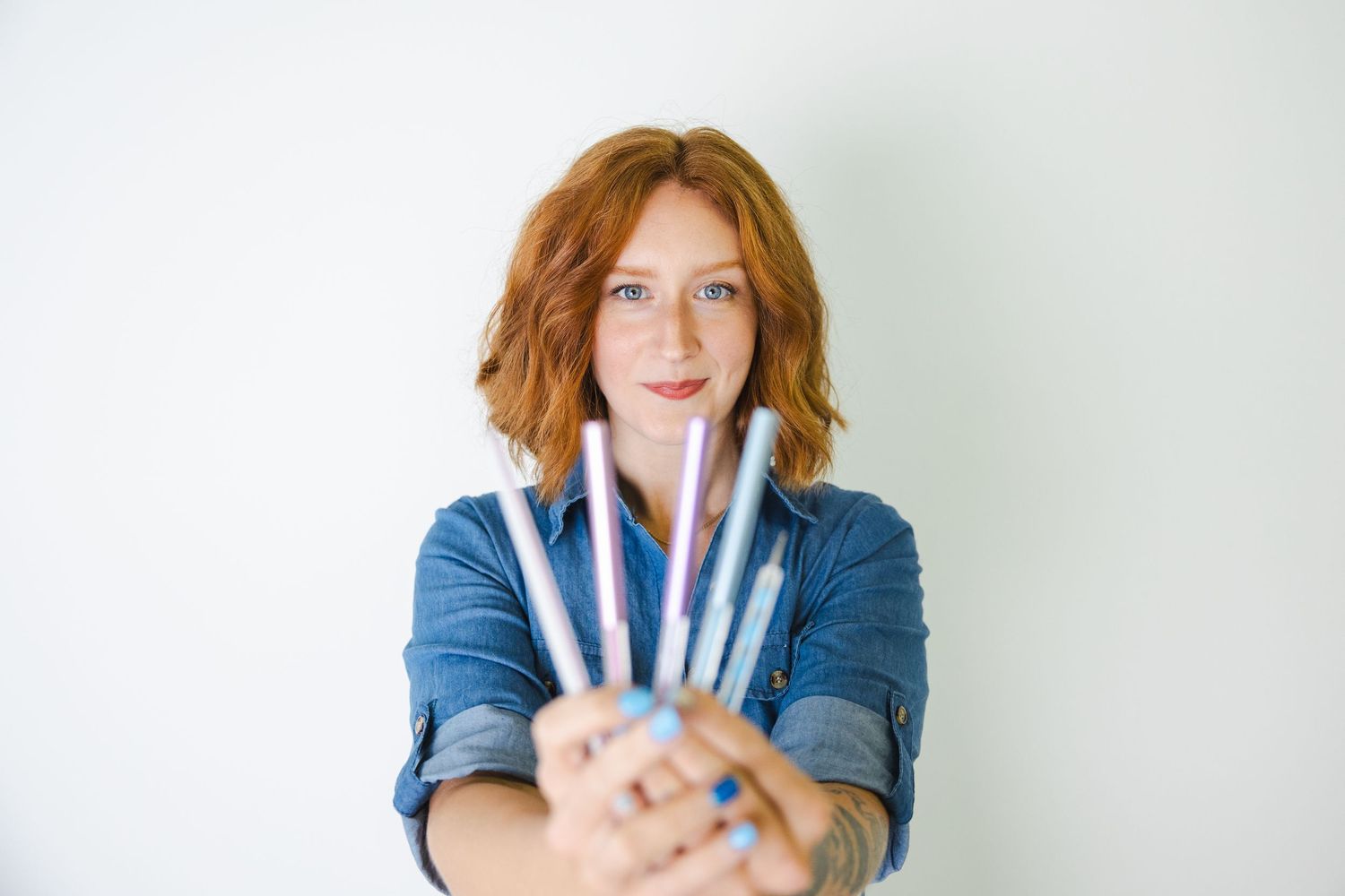 Person in denim jacket displays collection of makeup brushes against white backdrop.