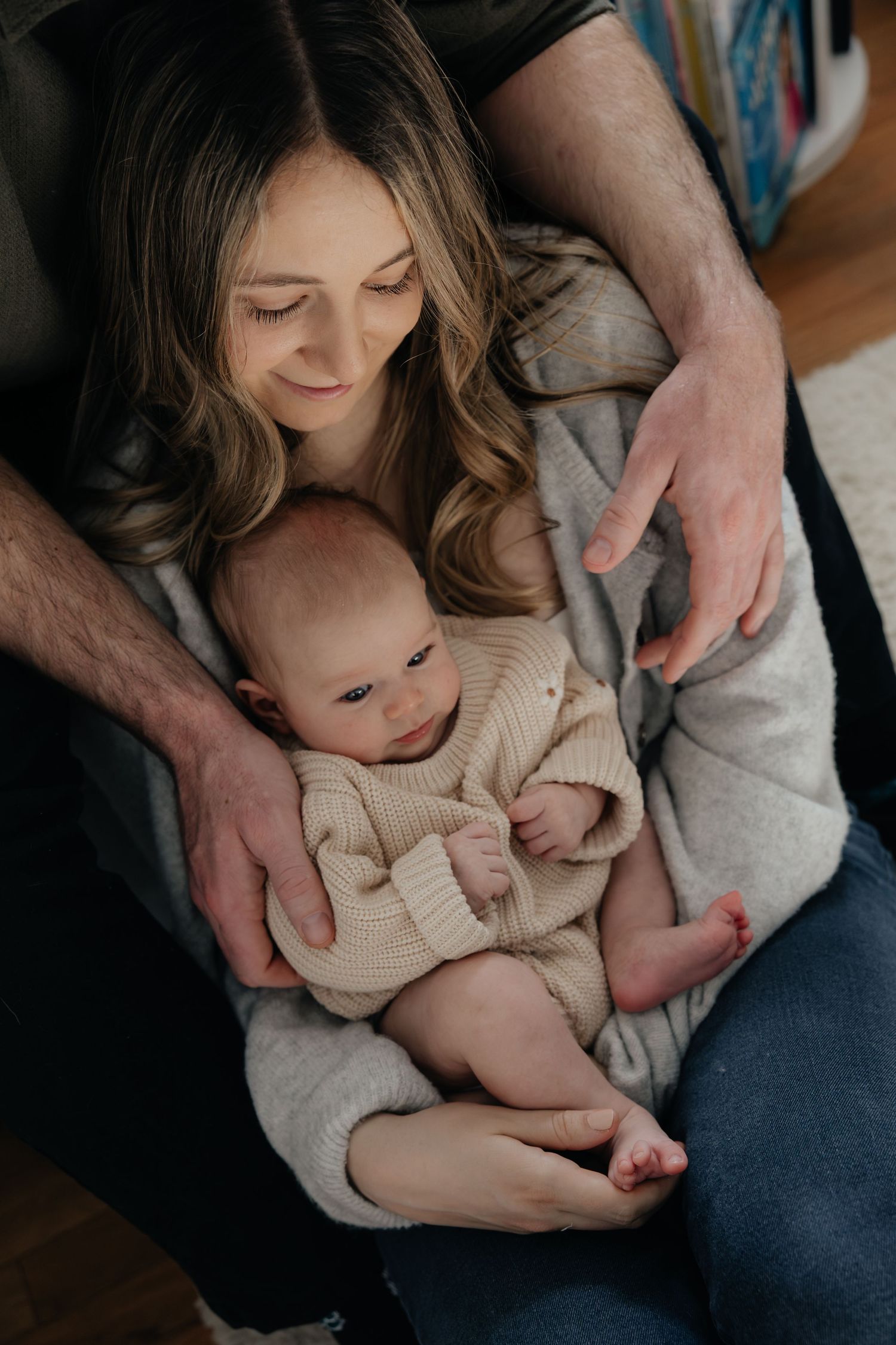 A mother tenderly holds her newborn baby while a father's hands gently support them both in a cozy home setting.