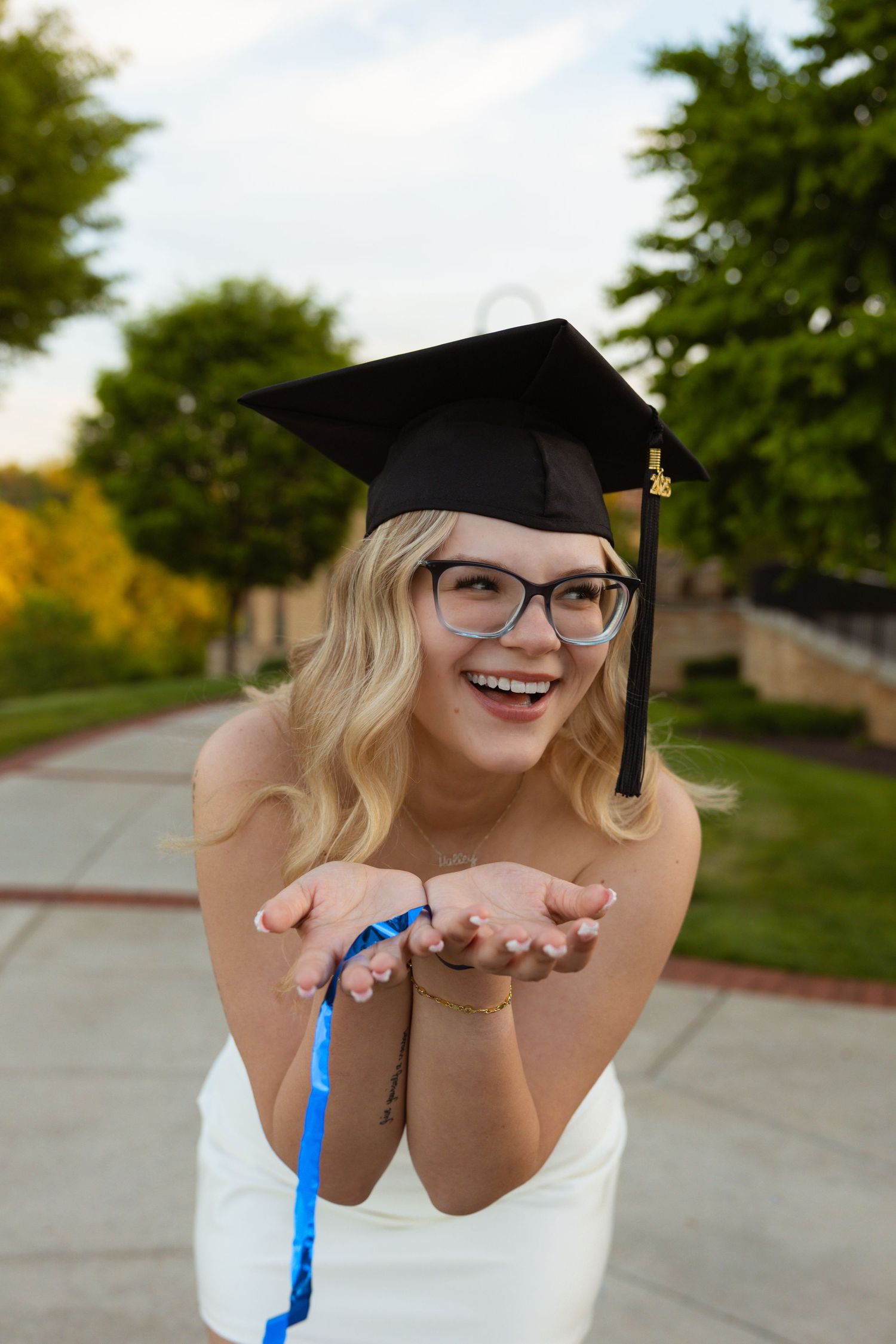 Graduate in cap and gown smiles while holding hands out during outdoor photoshoot.