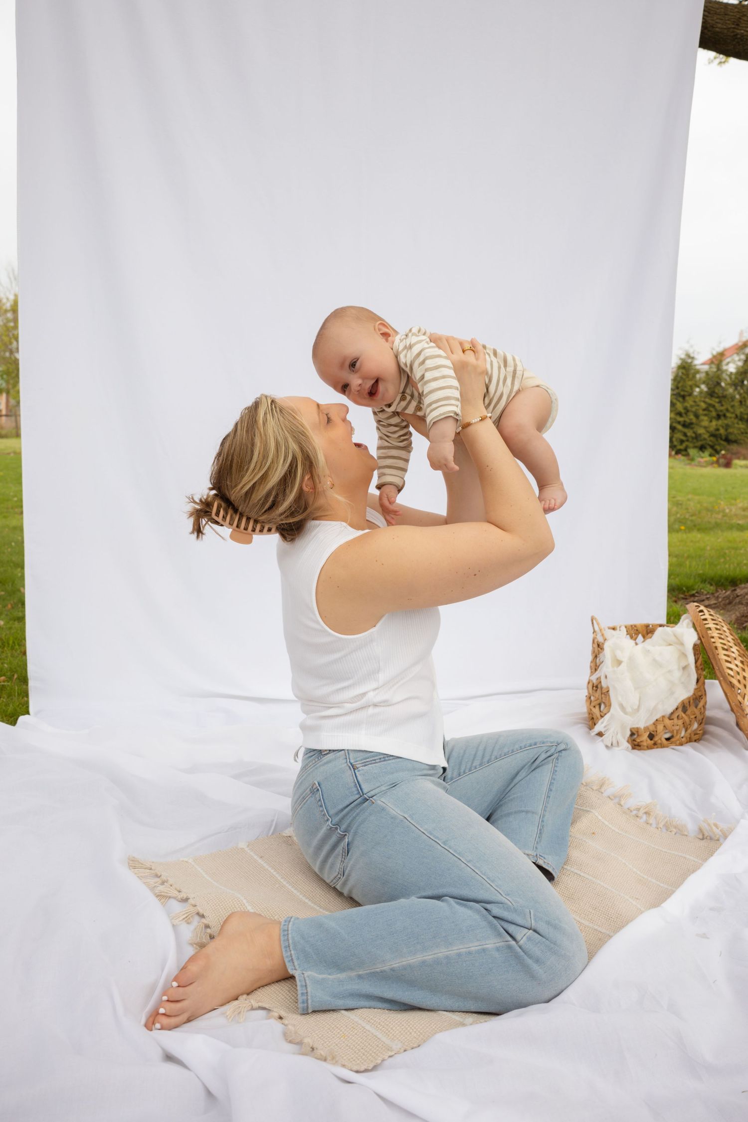 Parent plays with infant on white blanket during outdoor photo session.